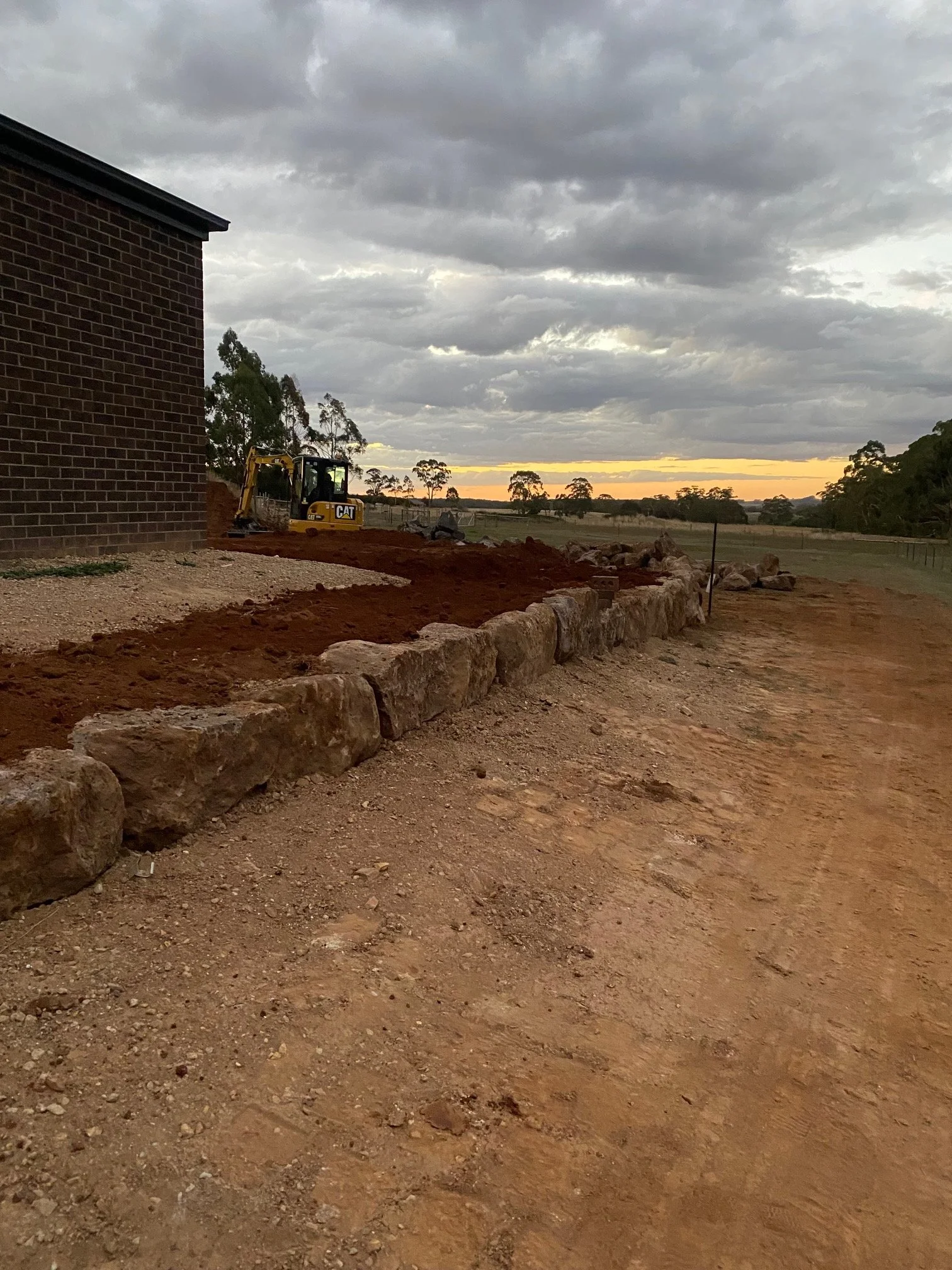 Construction site with dirt road and a small excavator near a brick building, with a cloudy sky and trees in the distance at sunset.