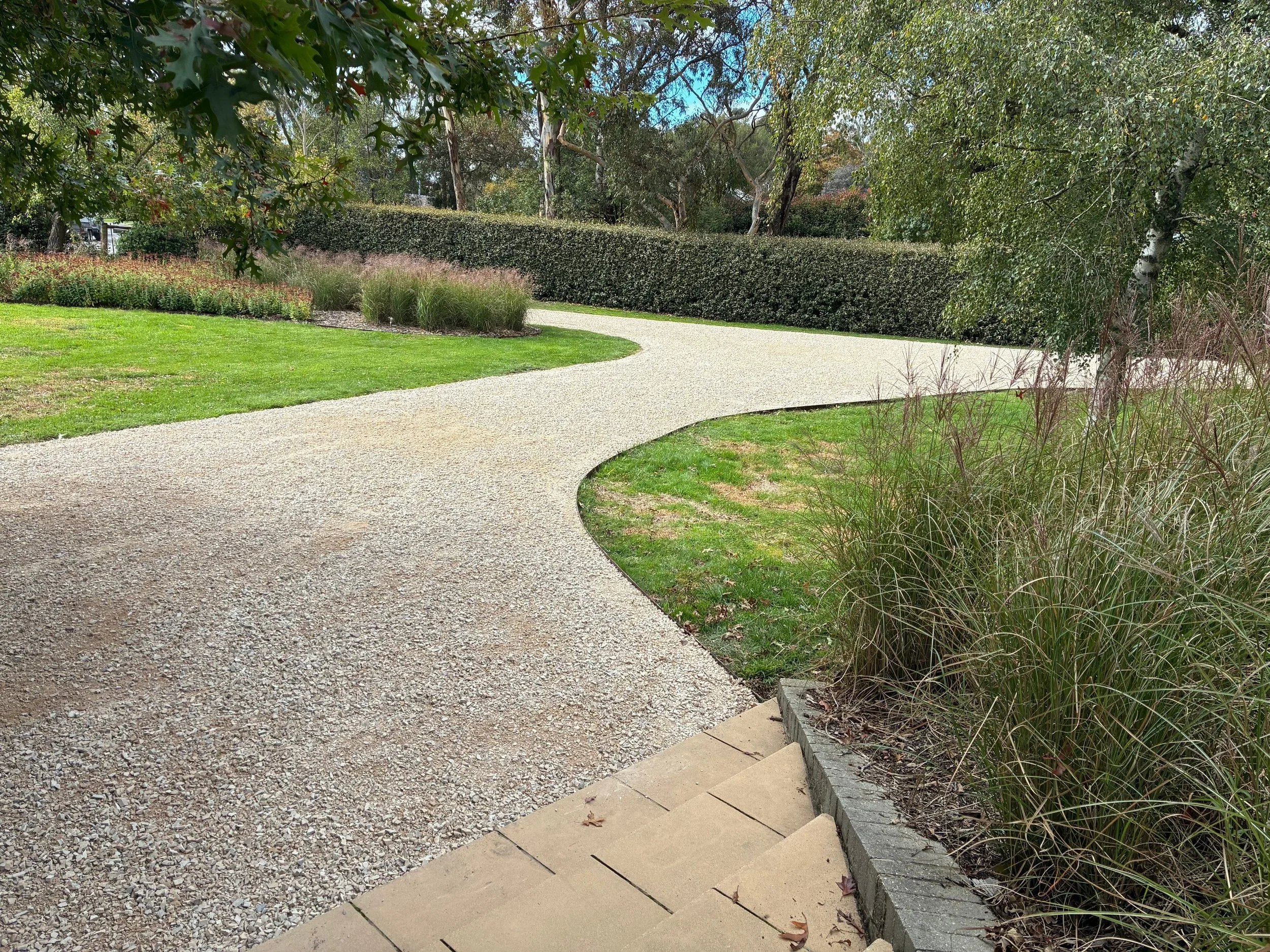 A winding gravel pathway in a well-maintained park or garden, bordered by green grass, bushes, and small trees, with taller trees and a hedge in the background.