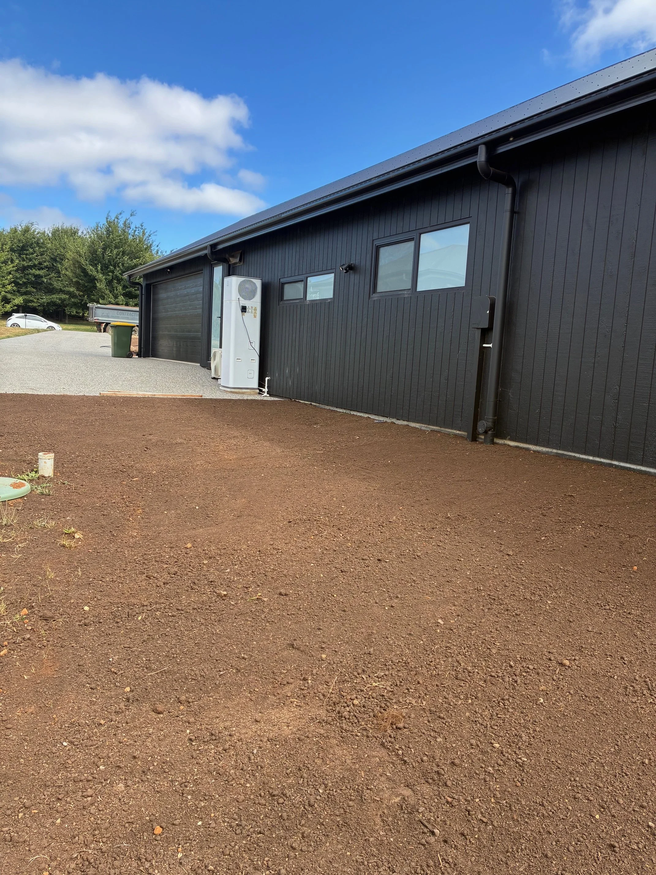 The backyard of a black house with a gray gravel driveway and a section of bare soil in the foreground. Two small windows, a door, and an air conditioning unit are visible on the house. A green trash bin and a parked car are in the background under a blue sky with scattered clouds.