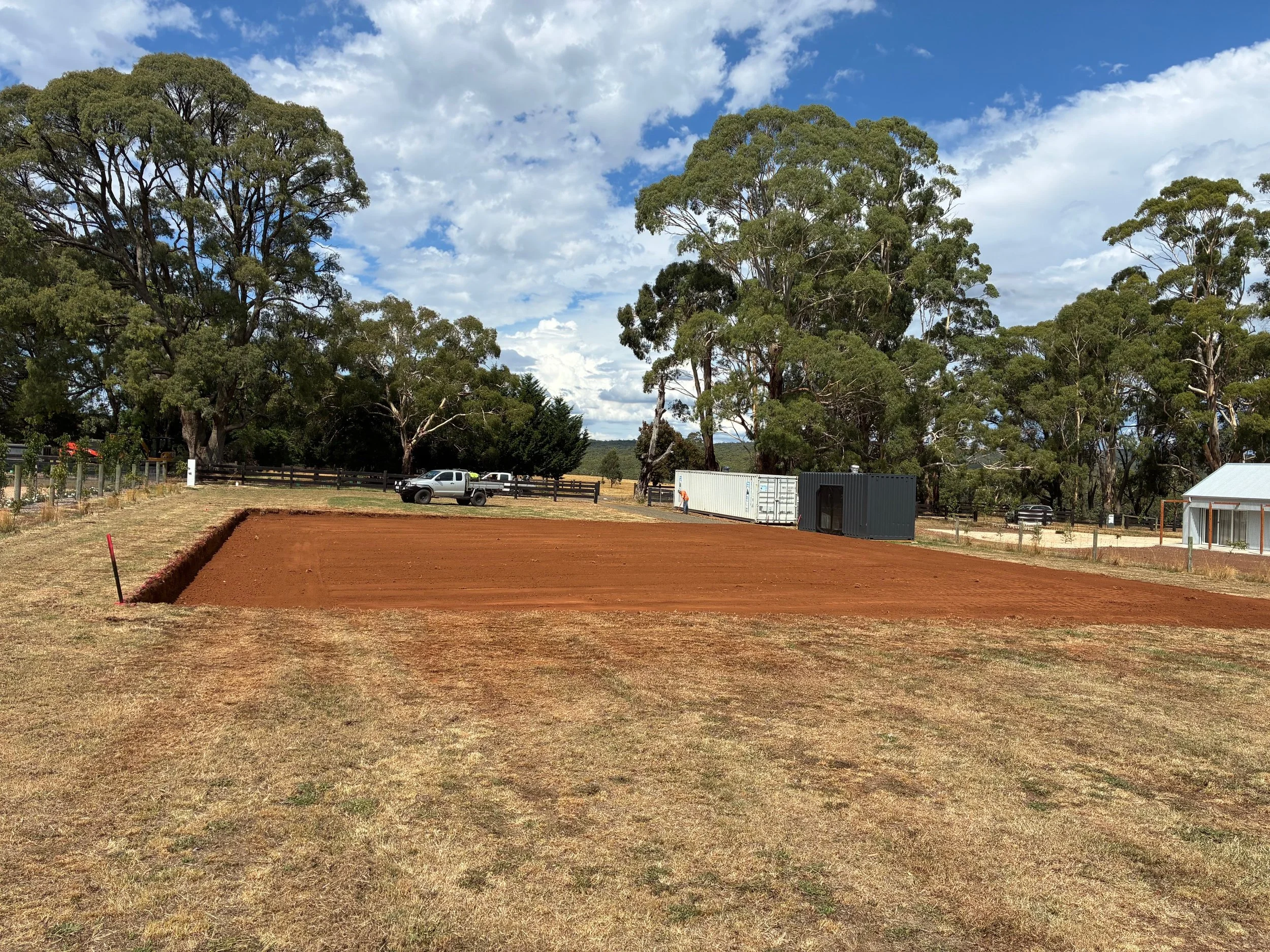 A sports field under construction with a layer of red dirt, surrounded by trees and a blue sky with clouds. There is a white and black storage container and vehicles parked in the background.