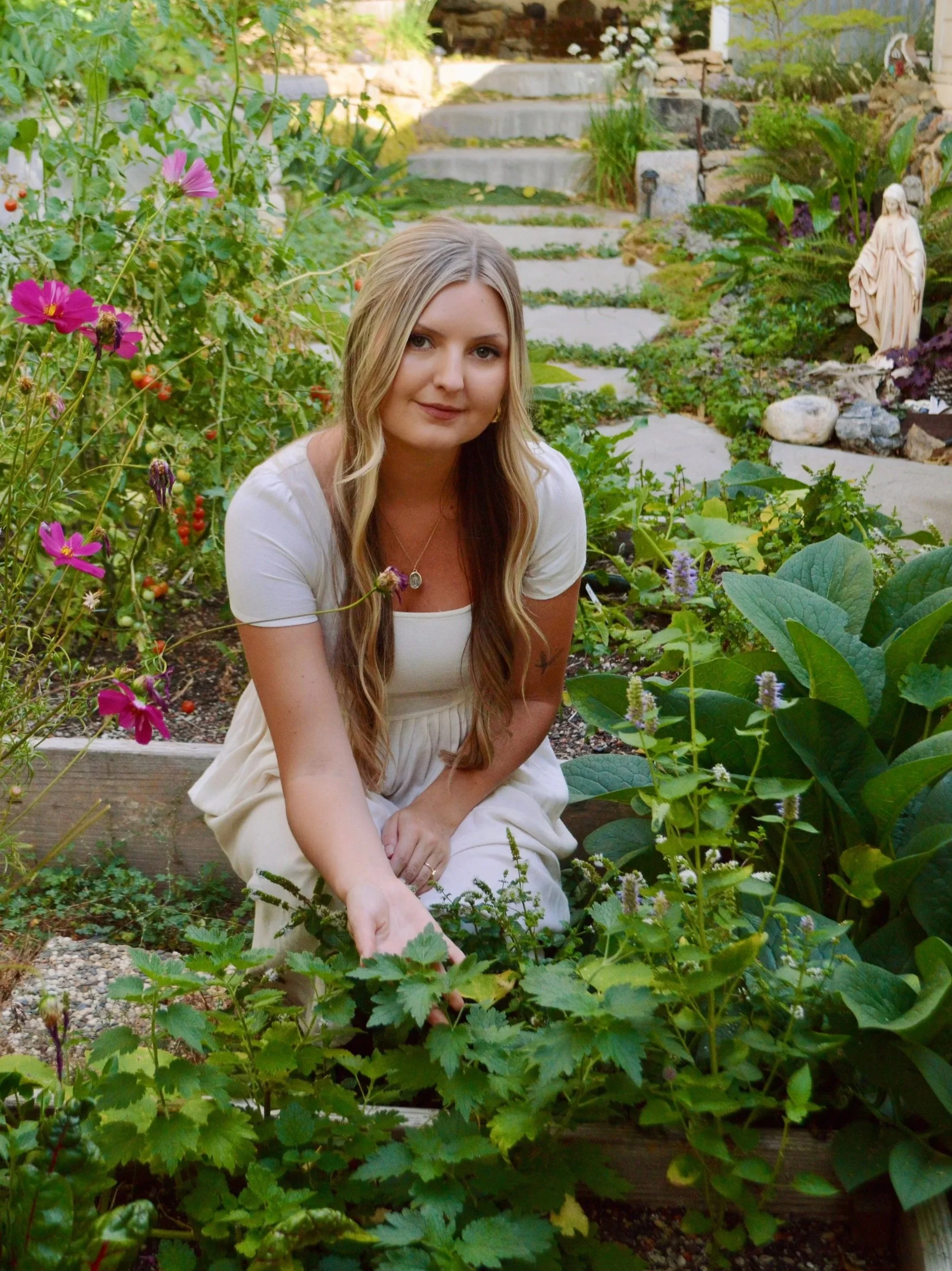 A woman with long blonde hair sitting in a garden surrounded by green plants and flowers, pointing to a plant with her finger.