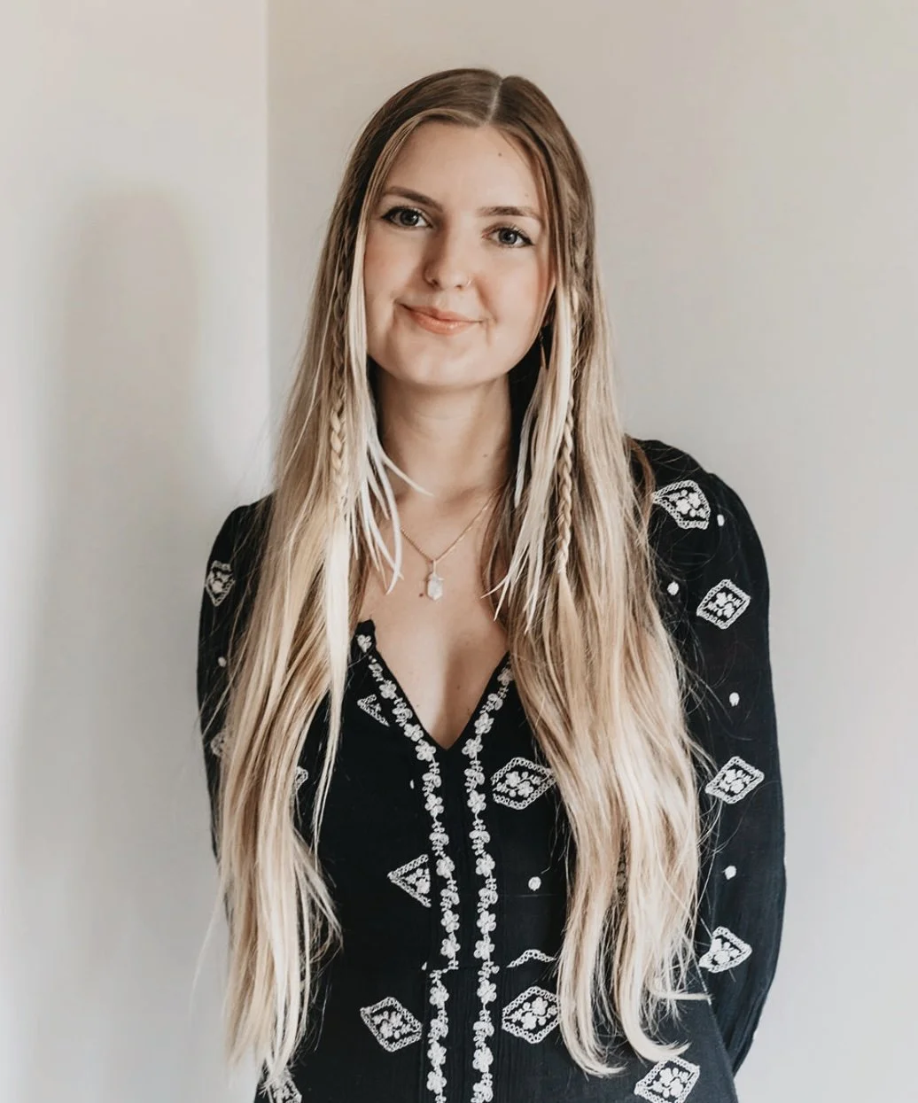 A woman with long, blonde hair and braided streaks, wearing a black dress with white decorative patterns, standing against a plain light-colored wall.