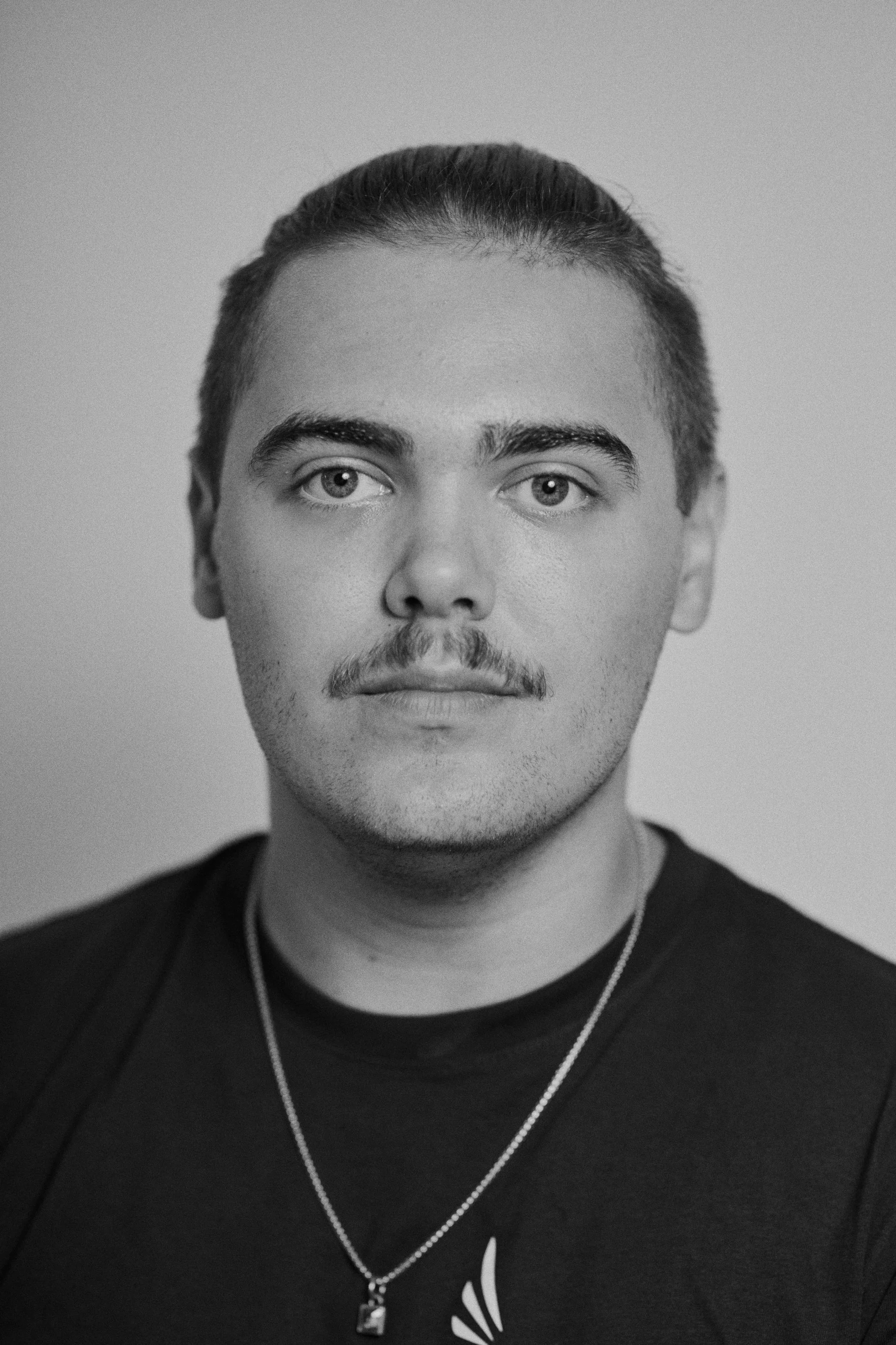 A black and white photo of a young man, River Bali, with tied bad dark hair, medium-light skin, and light-colored eyes. He is wearing a black shirt and a necklace. The background is plain and out of focus.