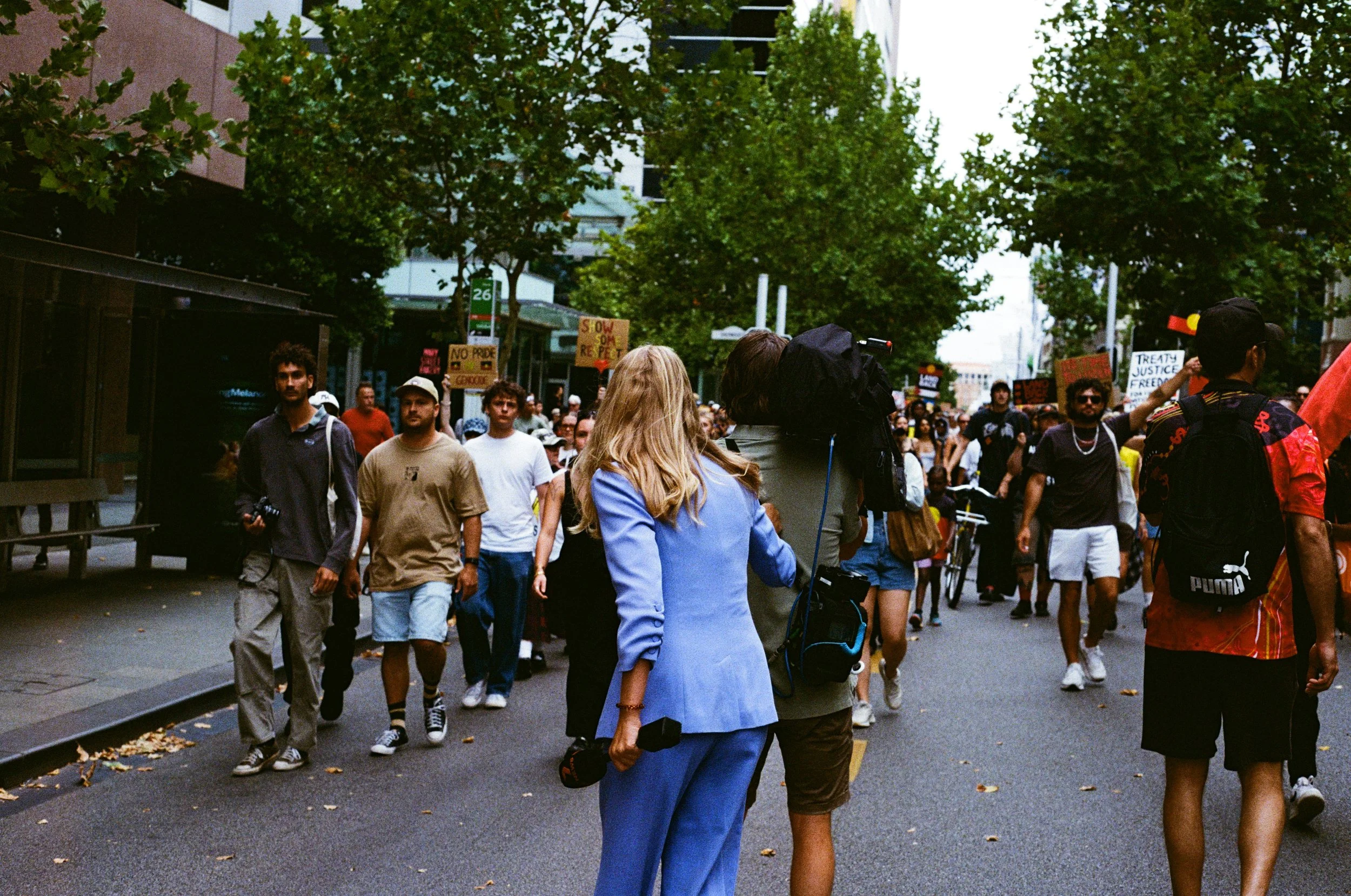 A diverse group of people participating in a street protest or march on a city street lined with trees and buildings, holding signs with messages about social justice.