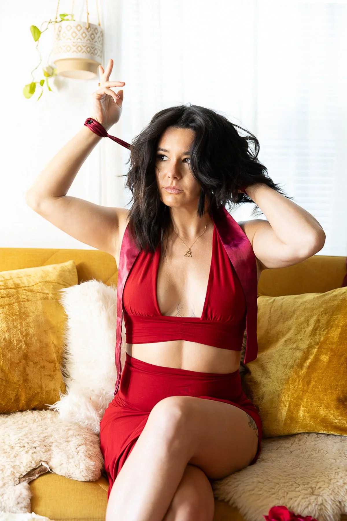 Woman with dark, toffee colored hair sitting on a yellow couch, wearing a red dress, adjusting her hair with one hand and making a peace sign with the other, surrounded by cushions and a hanging plant in a bright room.