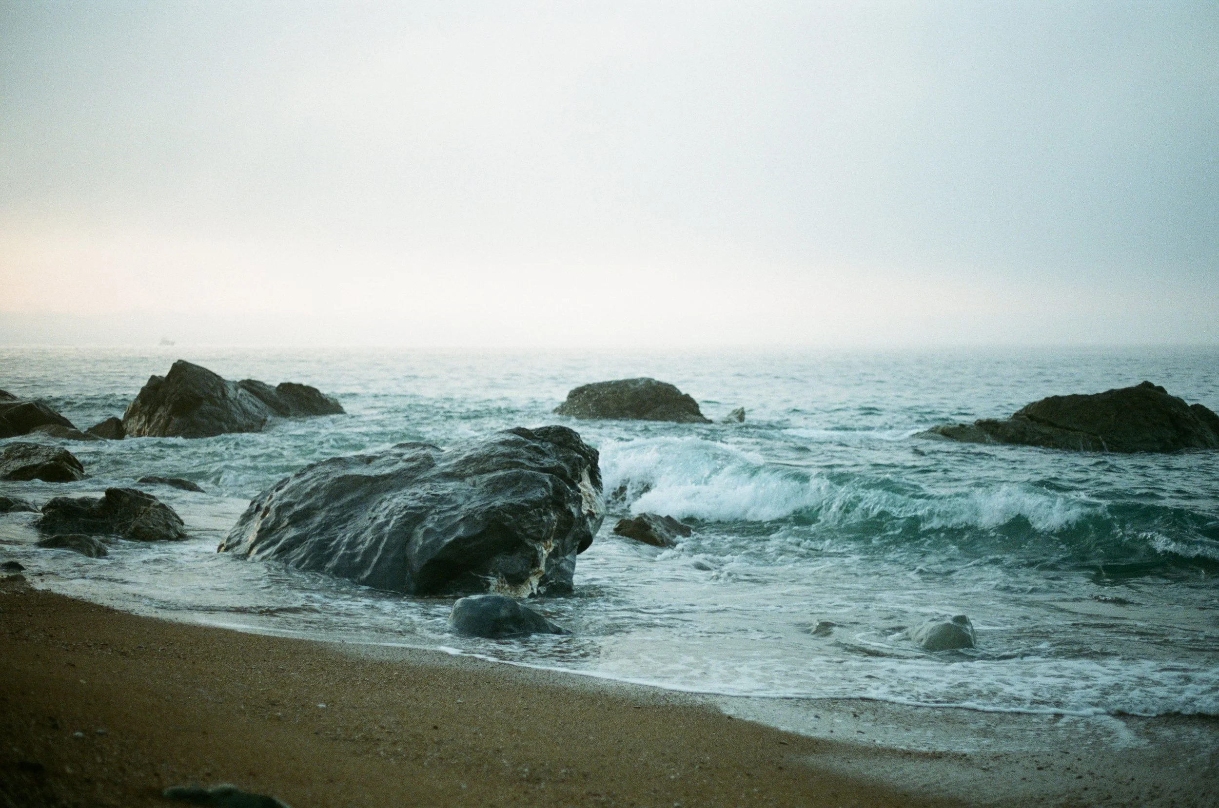 A beach scene with brown sand, large rocks in the water, and ocean waves crashing, under an overcast sky.
