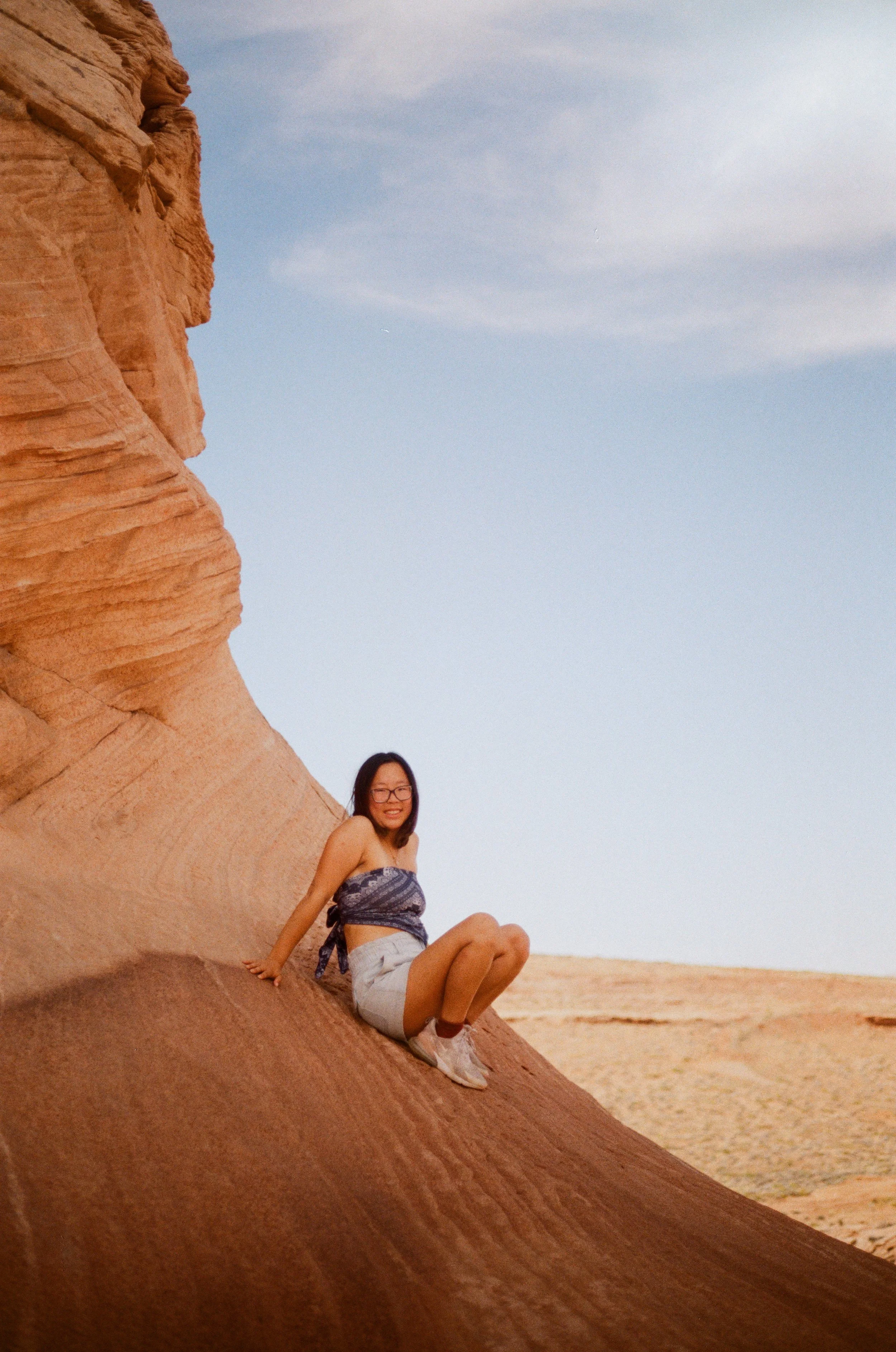A smiling woman in glasses, a blue patterned crop top, denim shorts, and sneakers, sitting on a large, smooth, reddish-brown rock formation in a desert landscape with a clear blue sky.
