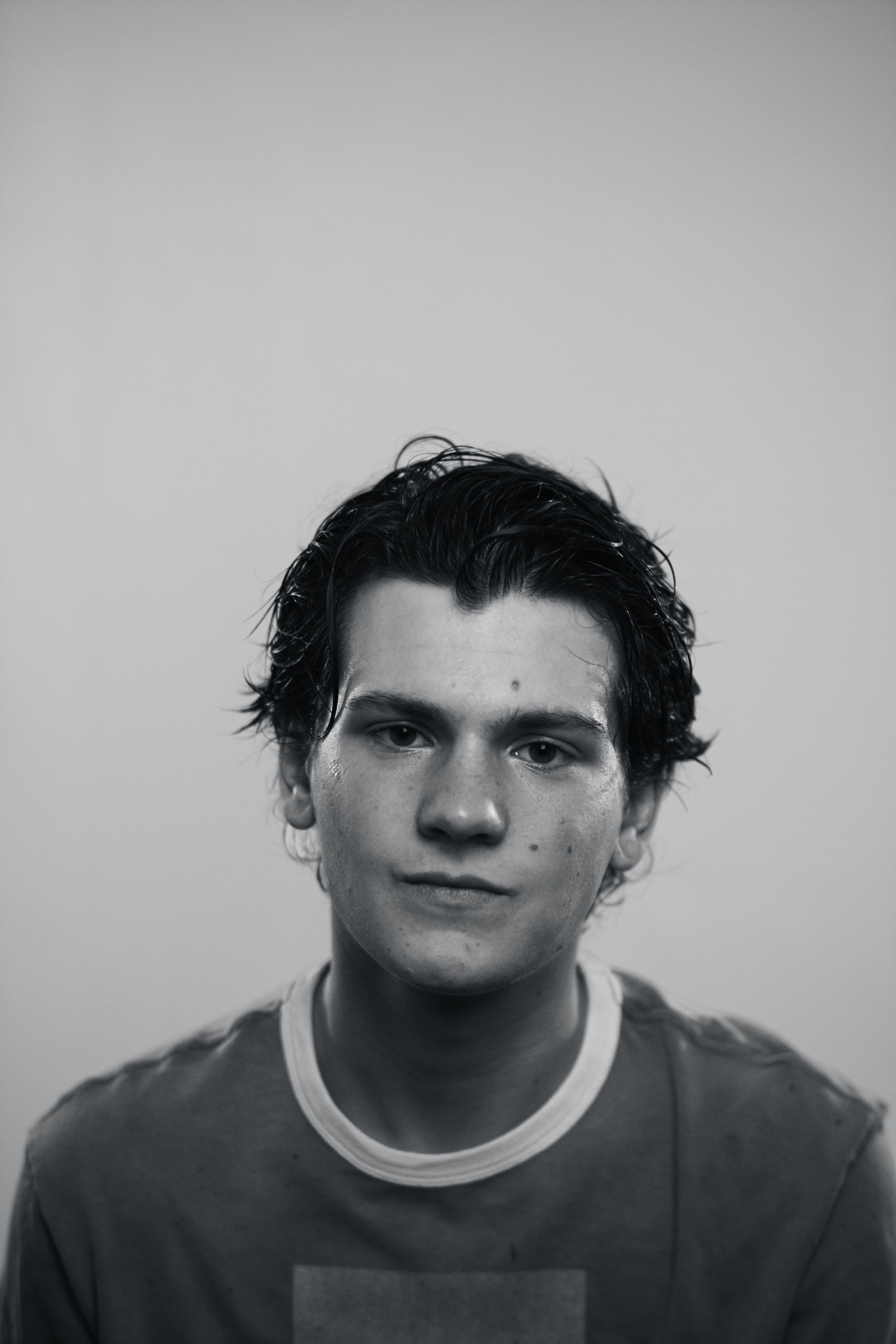 Black and white portrait of a young man with wet, tousled hair, wearing a t-shirt with a round neckline, smiling softly.
