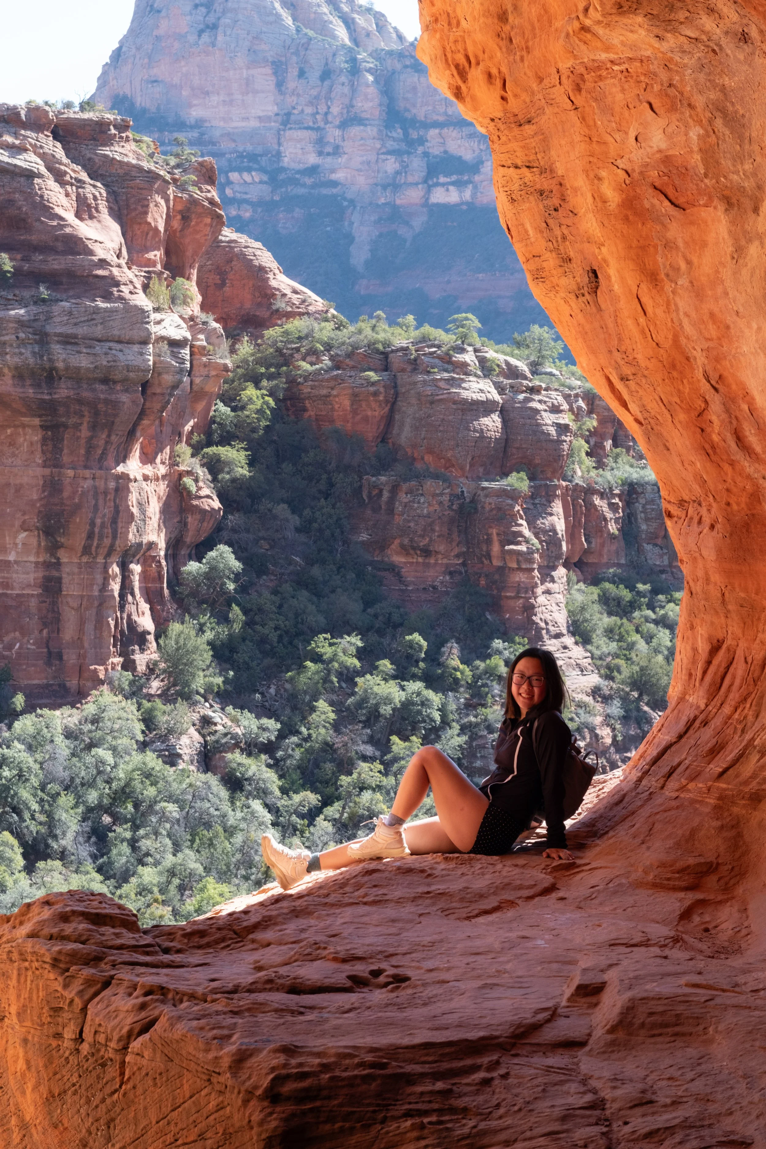 A young woman with glasses, black jacket, and shorts, sitting on a red rock ledge in a canyon, smiling at the camera. The background features towering red rock cliffs and lush green vegetation.