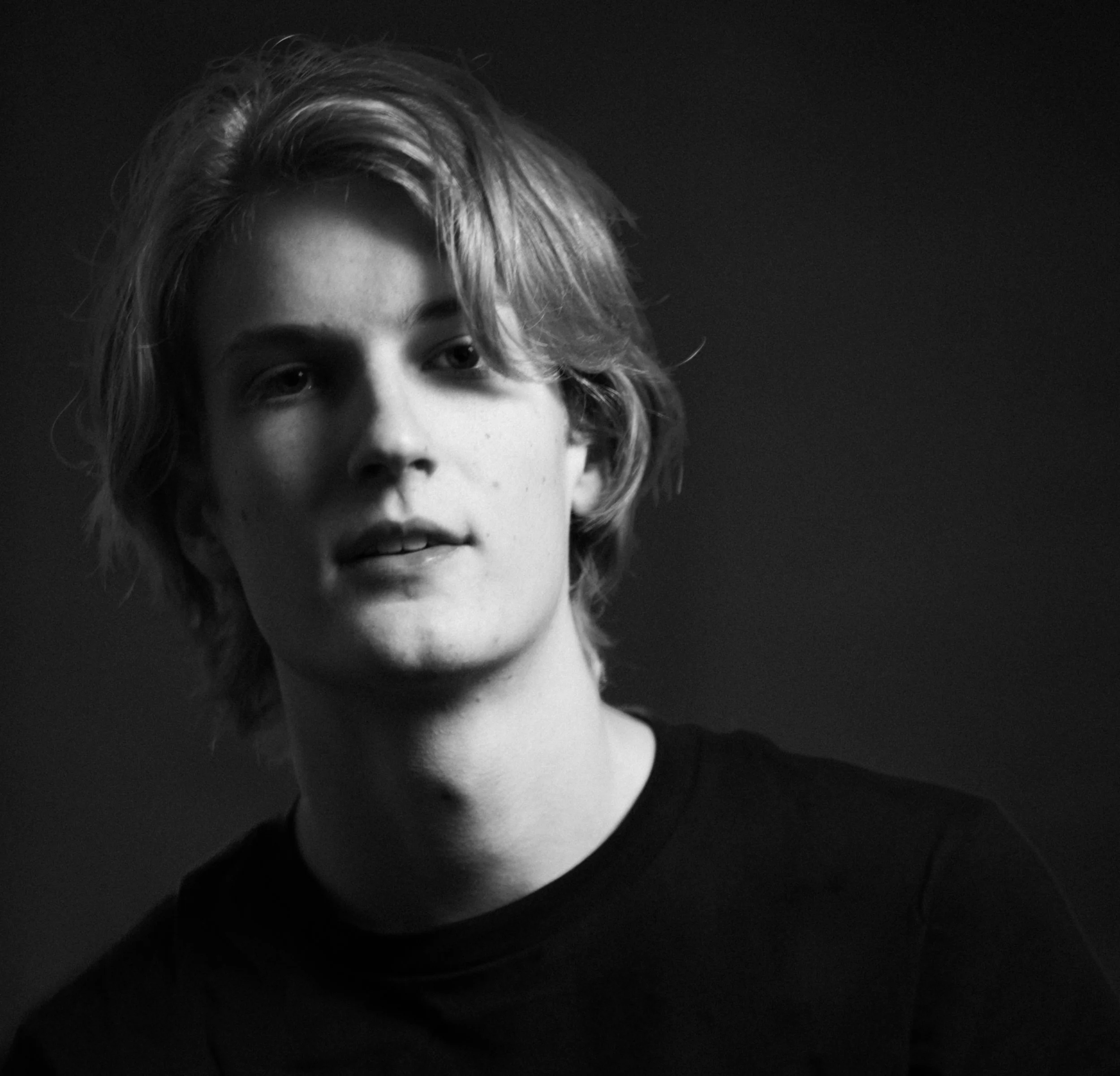 Black and white portrait of a young man with wavy, shoulder-length hair, wearing a dark shirt, looking slightly to the side.