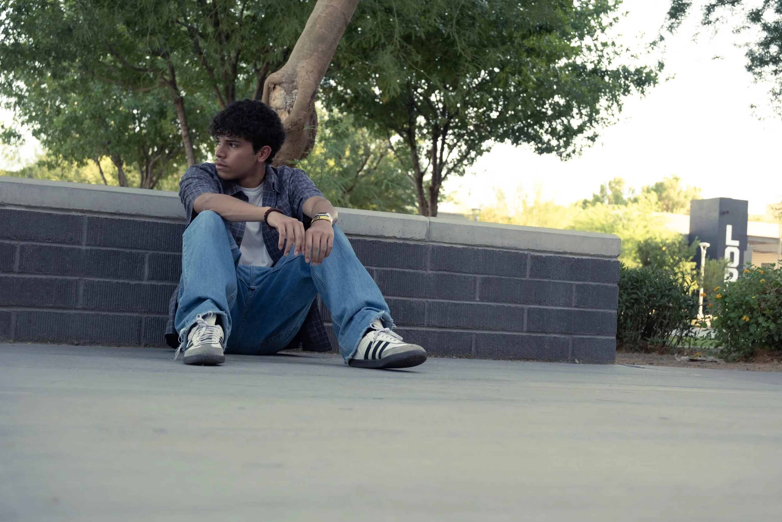 Young man sitting on the ground leaning against a brick wall, looking pensively to the side with trees and a building in the background.