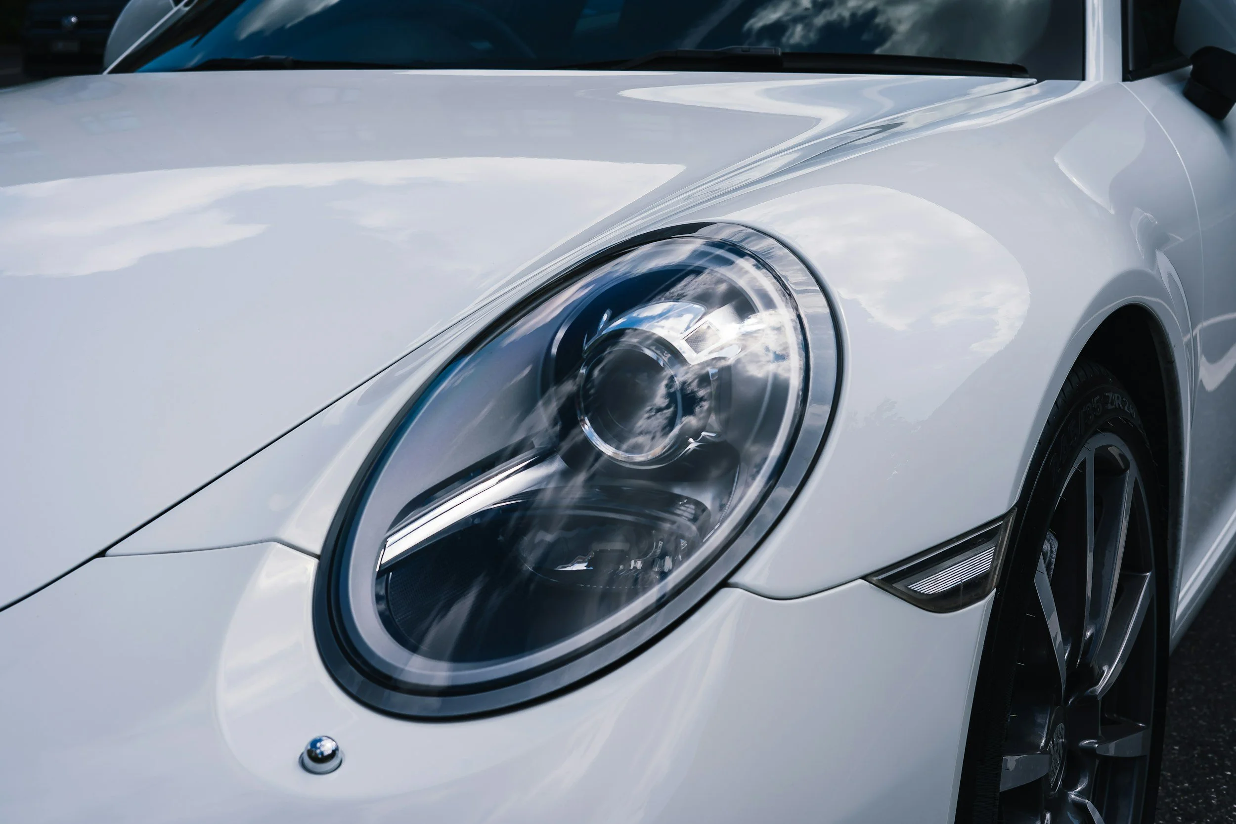 Close-up of the front left side of a white sports car, showing the large, rounded headlight and part of the hood and wheel.