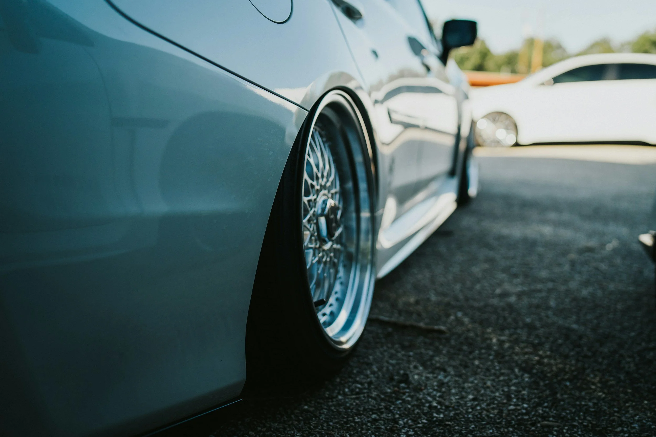 Close-up of a lowered sports car with custom wire wheels parked on asphalt, with another white car in the background.