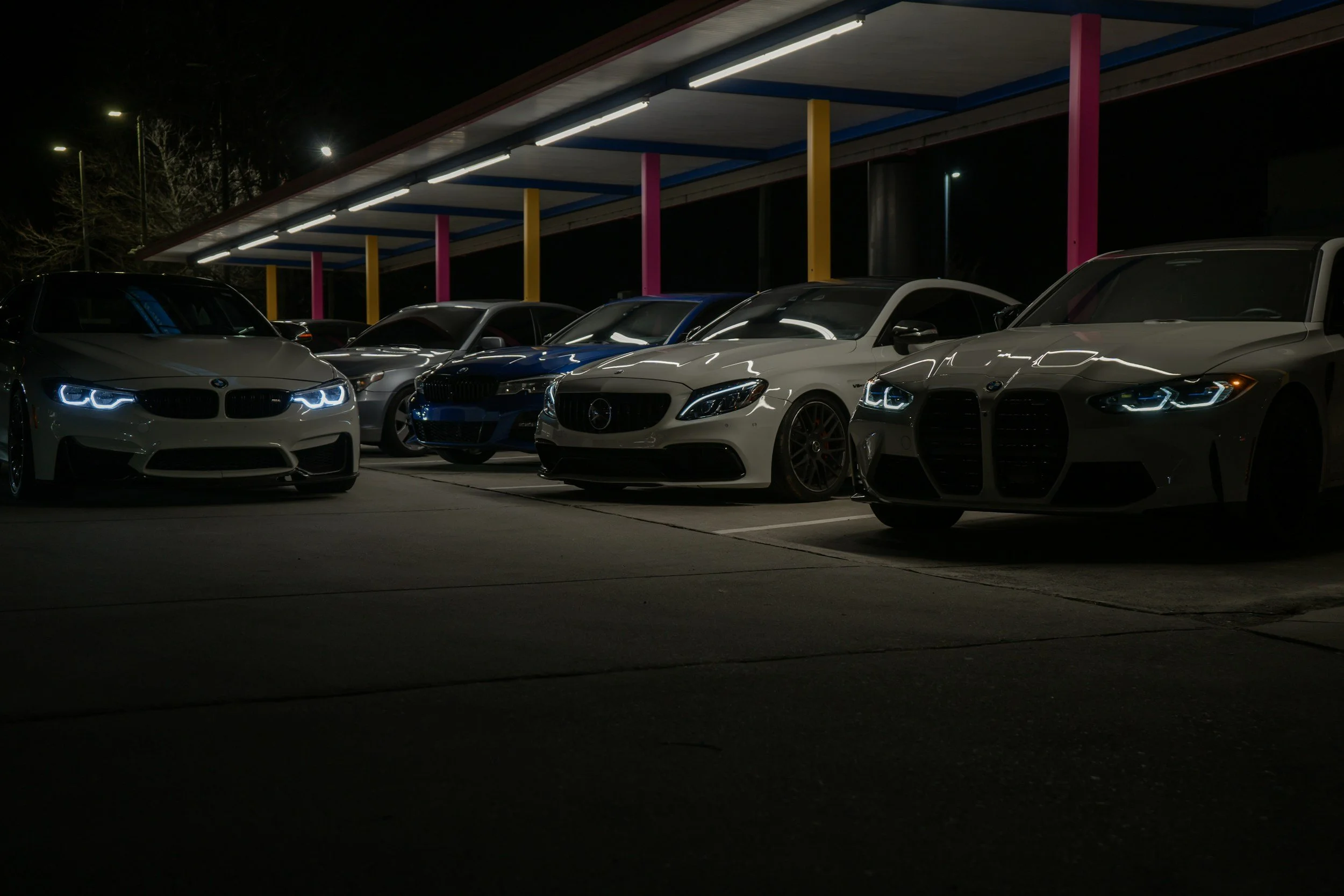 Nighttime view of a parking lot with five parked cars, including a BMW, a blue car, and an Alfa Romeo, under a colorful covered structure with pink, yellow, and blue posts.
