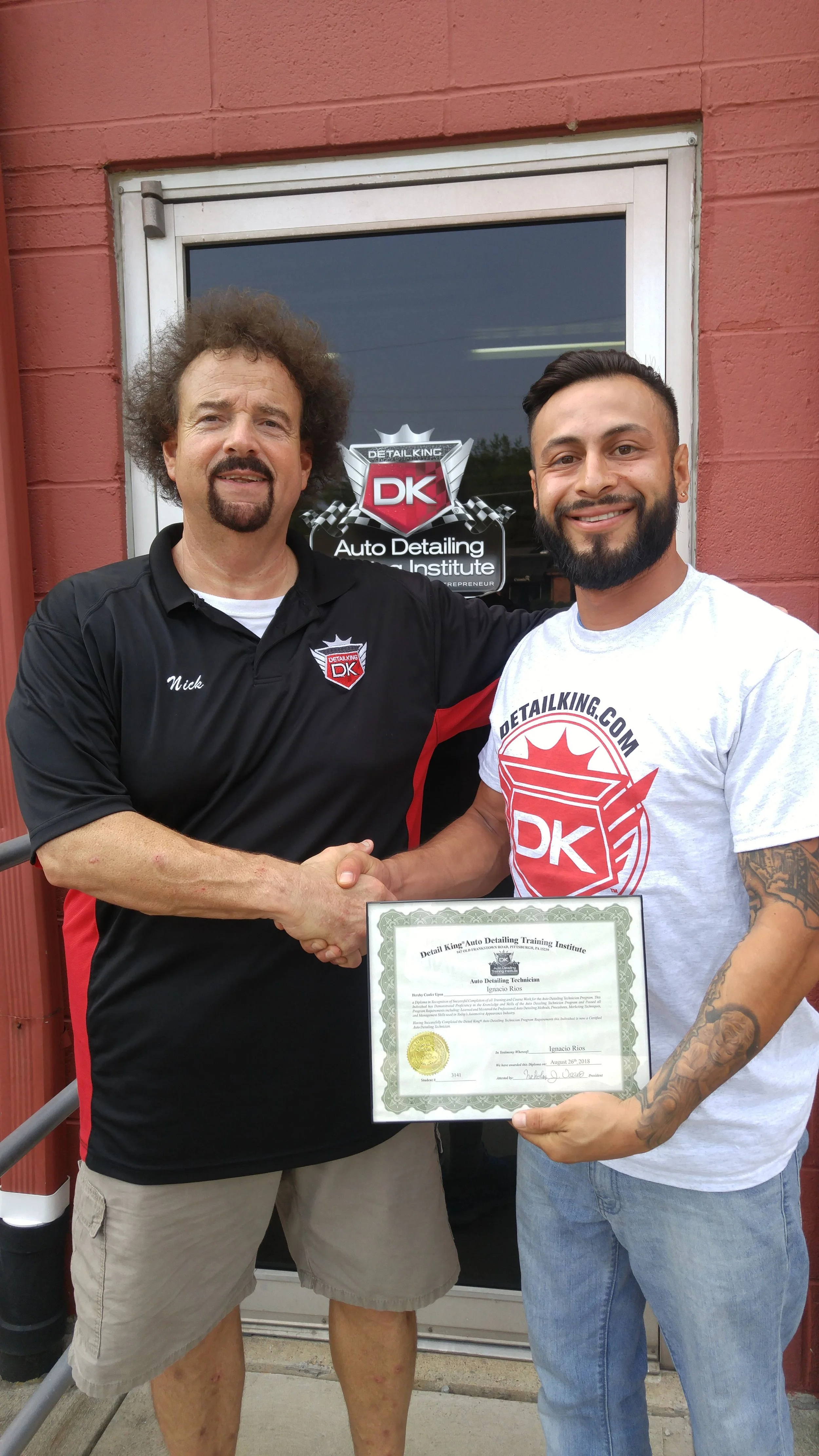 Two men shaking hands, one man is holding a framed certificate. They are outside a building with a sign behind them that reads "Auto Detailing Training Institute" and has a logo with "DK" on it.