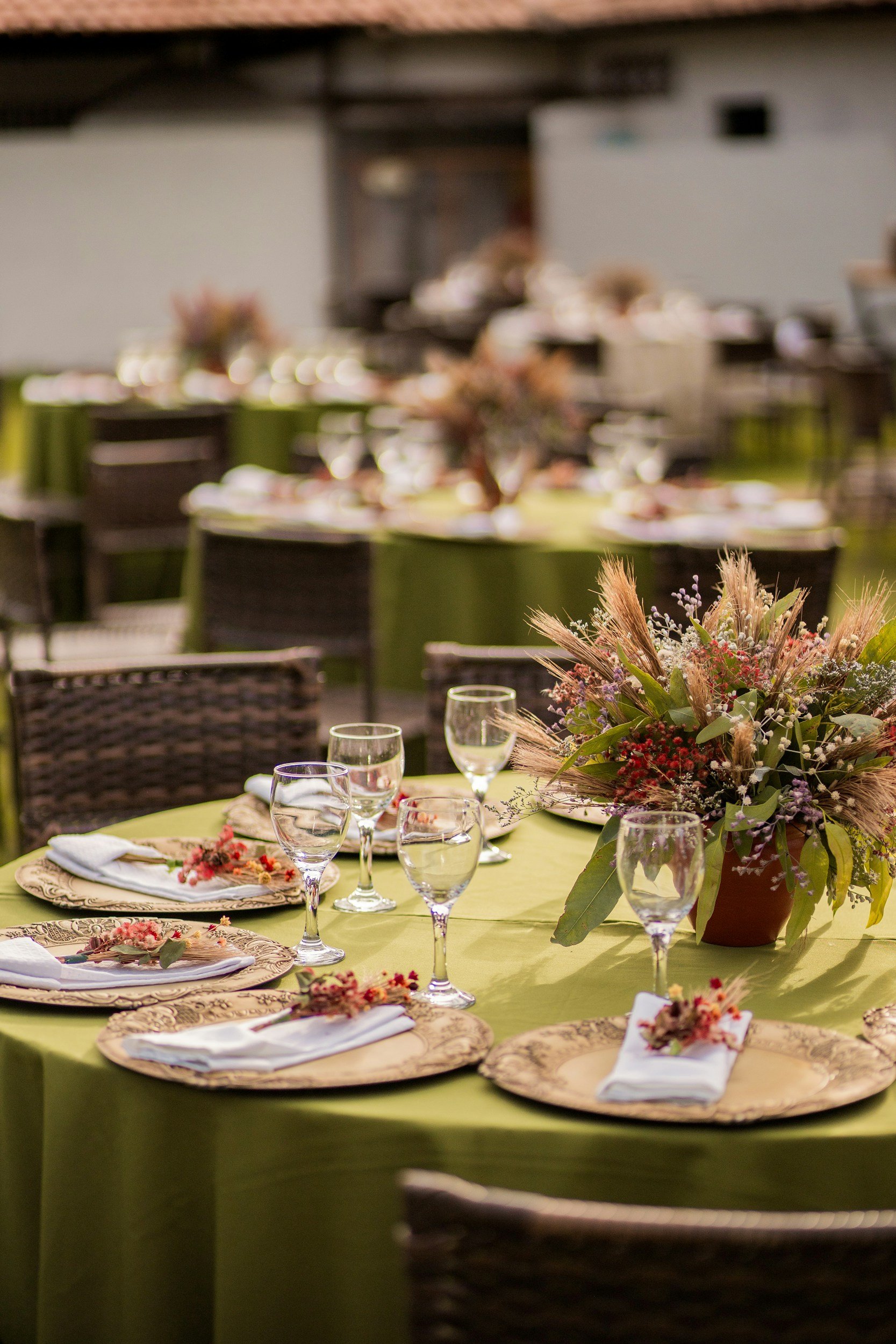 Table set for an event with a green tablecloth, decorative plates, glasses, cloth napkins with small floral arrangements, and a large centerpiece with flowers and dried grasses.