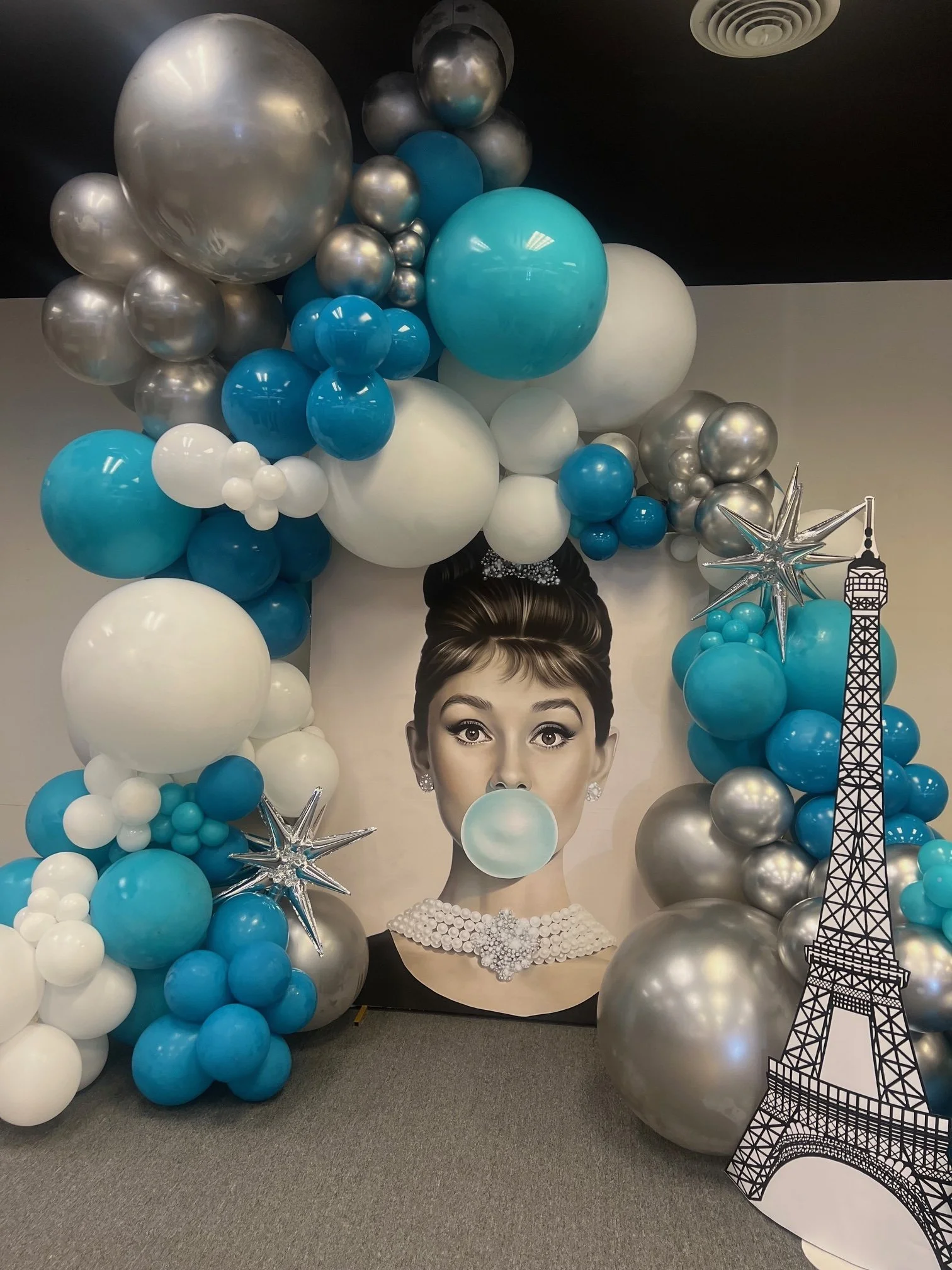 Decorative balloon arrangement in blue, white, and silver surrounding a large portrait of Audrey Hepburn blowing a bubble with gum, with Eiffel Tower and star-shaped balloons.