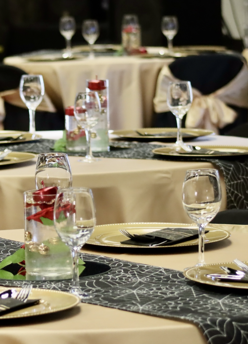 Elegant banquet table setup with wine glasses, water glasses, gold chargers, black napkins, and a black table runner with spiderwebs, decorated with red flowers and green leaves.