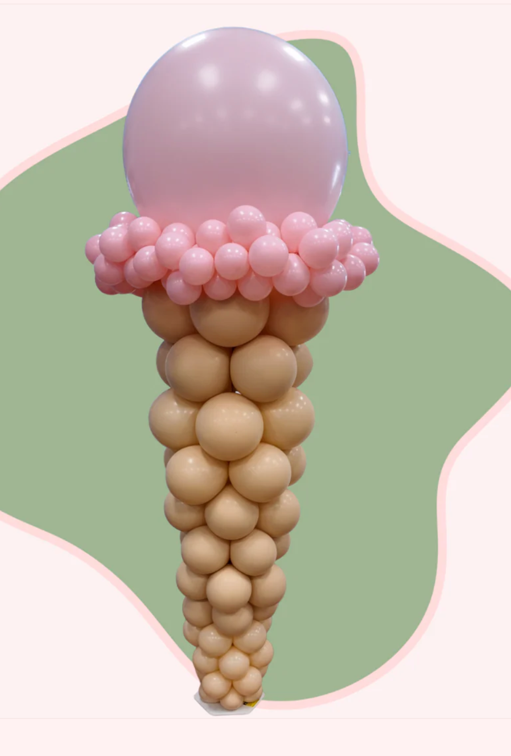 Balloon sculpture resembling an ice cream cone with a pink top scoop, a middle pink decorative layer, and a beige cone, set against a pastel green and pink background.