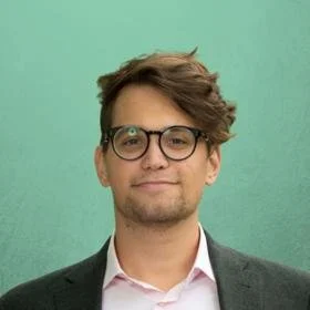 A man with brown hair, glasses, and a slight smile, wearing a dark blazer over a pink shirt, standing in front of a mint green background.