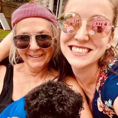 Two women smiling outdoors with a black dog, wearing sunglasses and casual clothing.