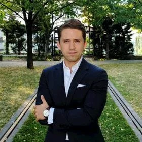 A man in a dark suit with a white shirt and pocket square standing outdoors on a pathway with trees in the background.
