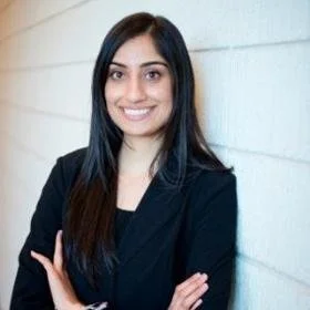 A woman with long dark hair, smiling, wearing a black blazer, standing against a white wall.