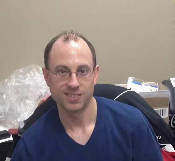 Portrait of a man with short dark hair, glasses, and a slight smile, wearing a blue shirt, sitting in an office or medical setting with various items in the background.
