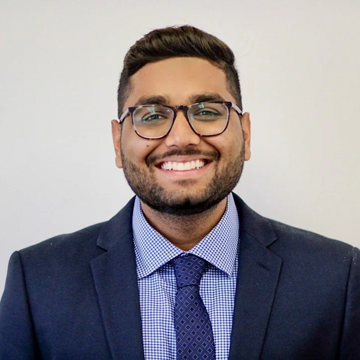 A smiling man with glasses, wearing a dark suit and tie, against a plain white background.