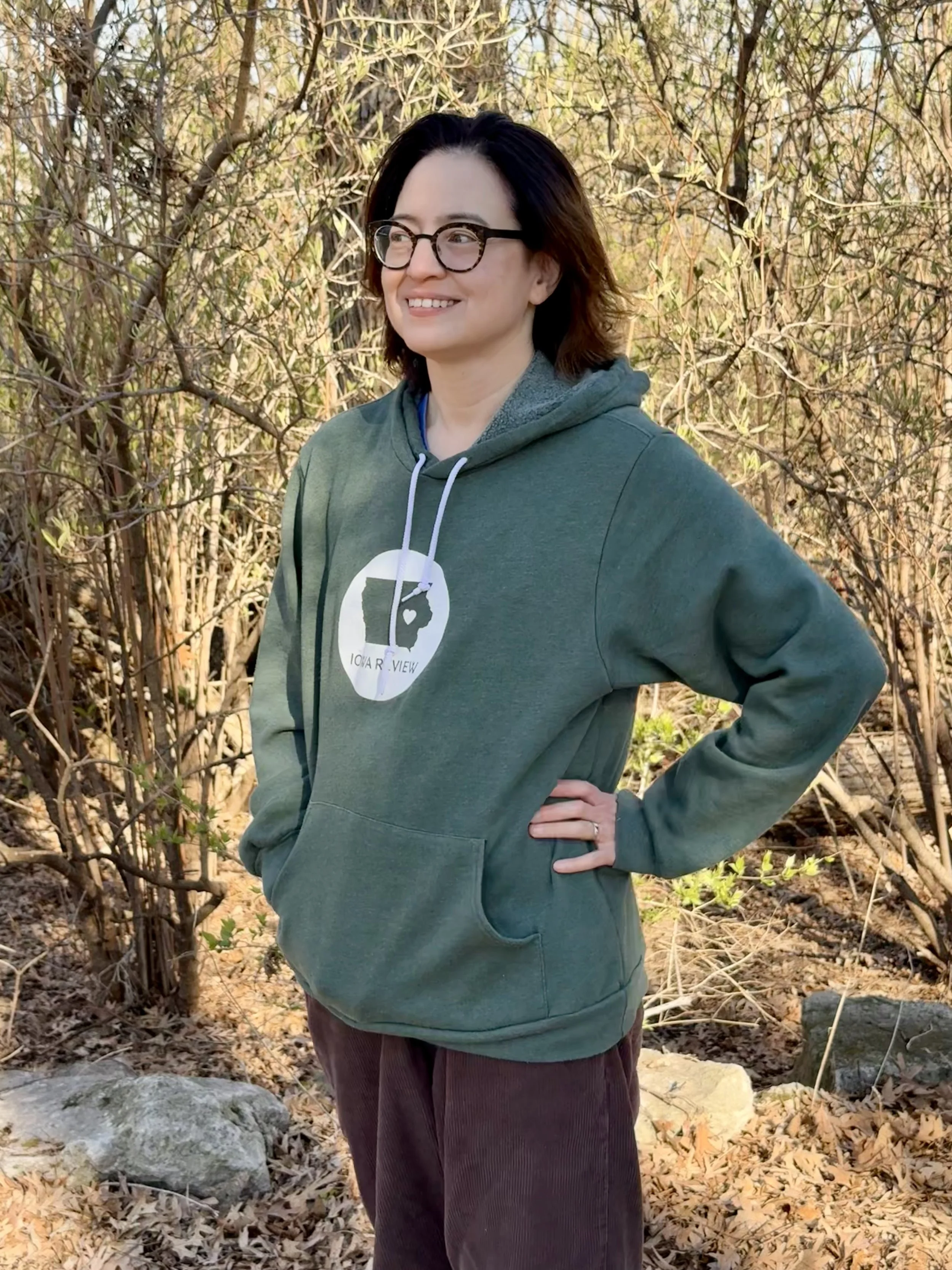 Lynne Nugent is standing outdoors in front of trees, wearing a green Iowa Review hoodie. She has her hand on her hip and is smiling.
