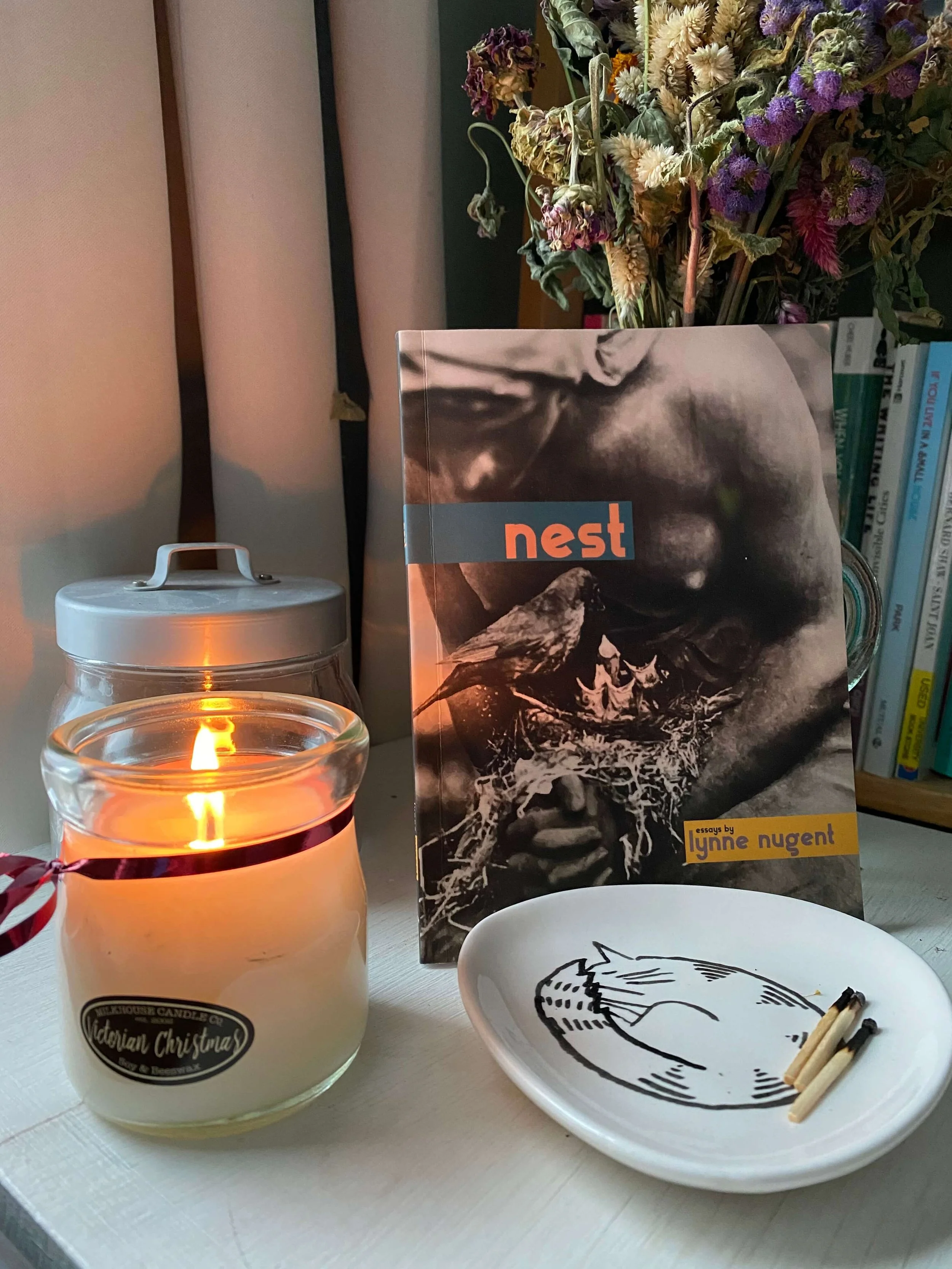 Lynne Nugent's chapbook, Nest, on a table with a lit candle, a white ceramic dish with a black minimalist illustration of a cat, and a bouquet of dried flowers in the background.