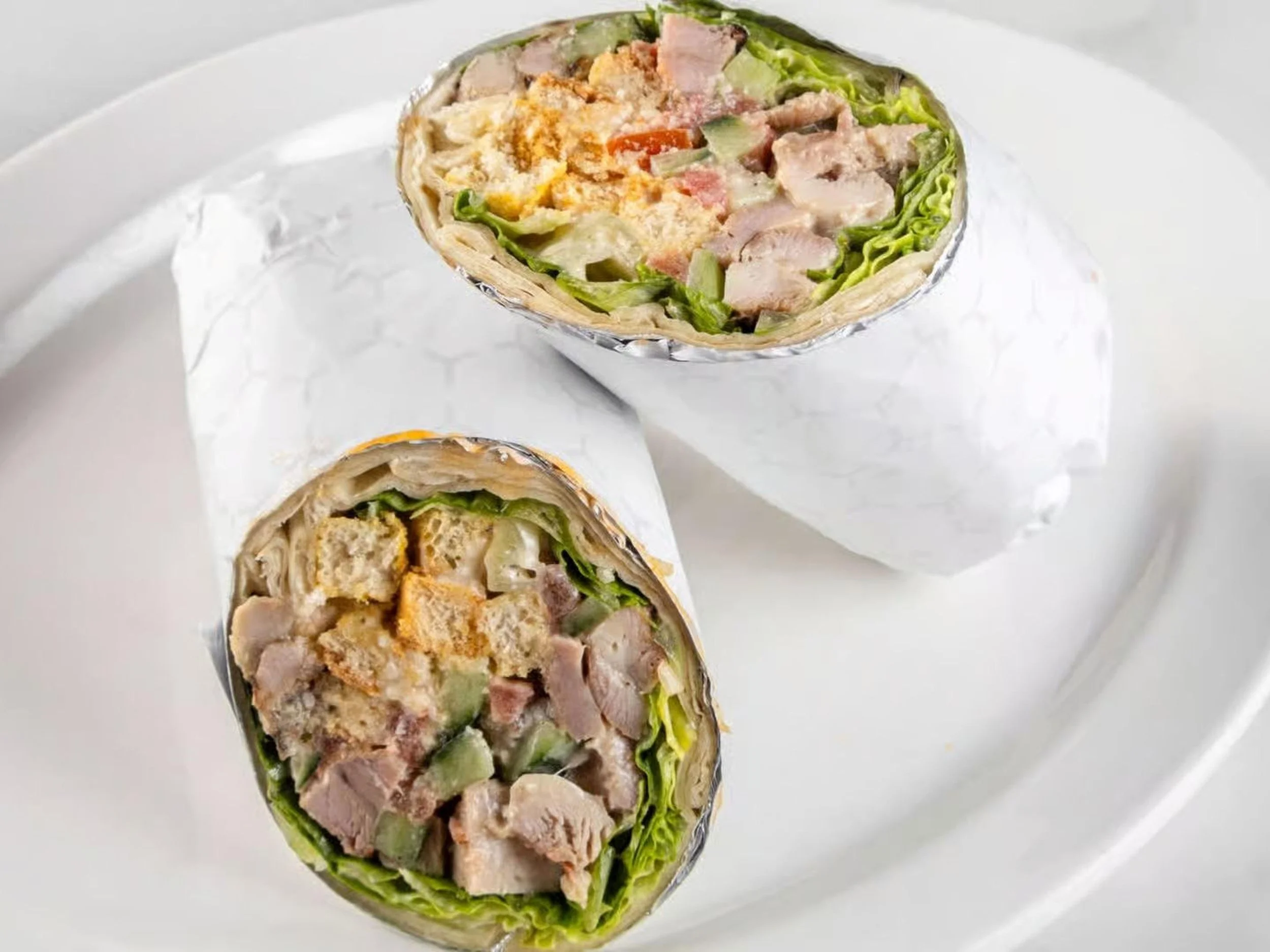 Two cut-in-half chicken Caesar wraps with lettuce, chicken pieces, and Caesar dressing on a white plate.