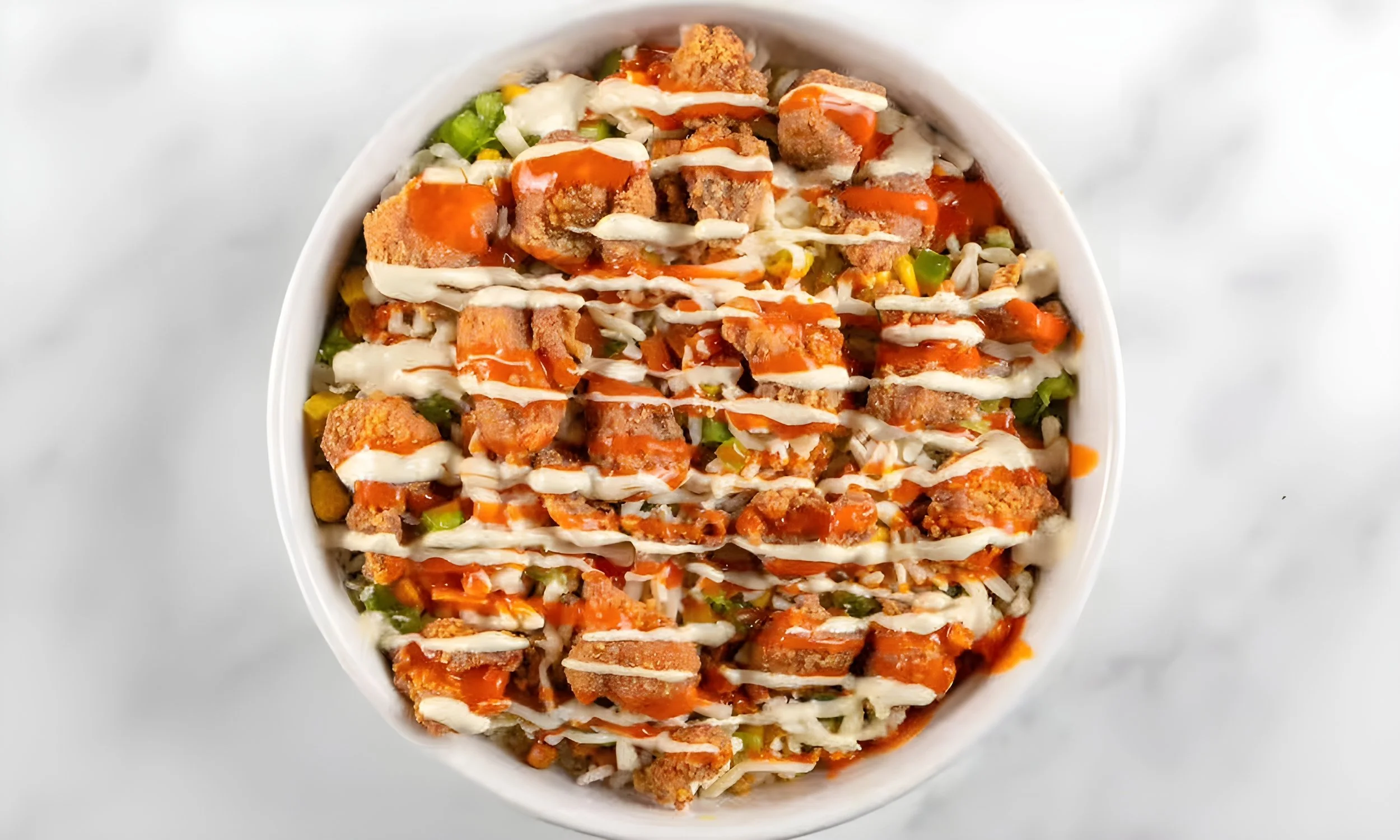Bowl of fried rice topped with mayonnaise, hot sauce, and fried chicken pieces surrounded by diced vegetables.