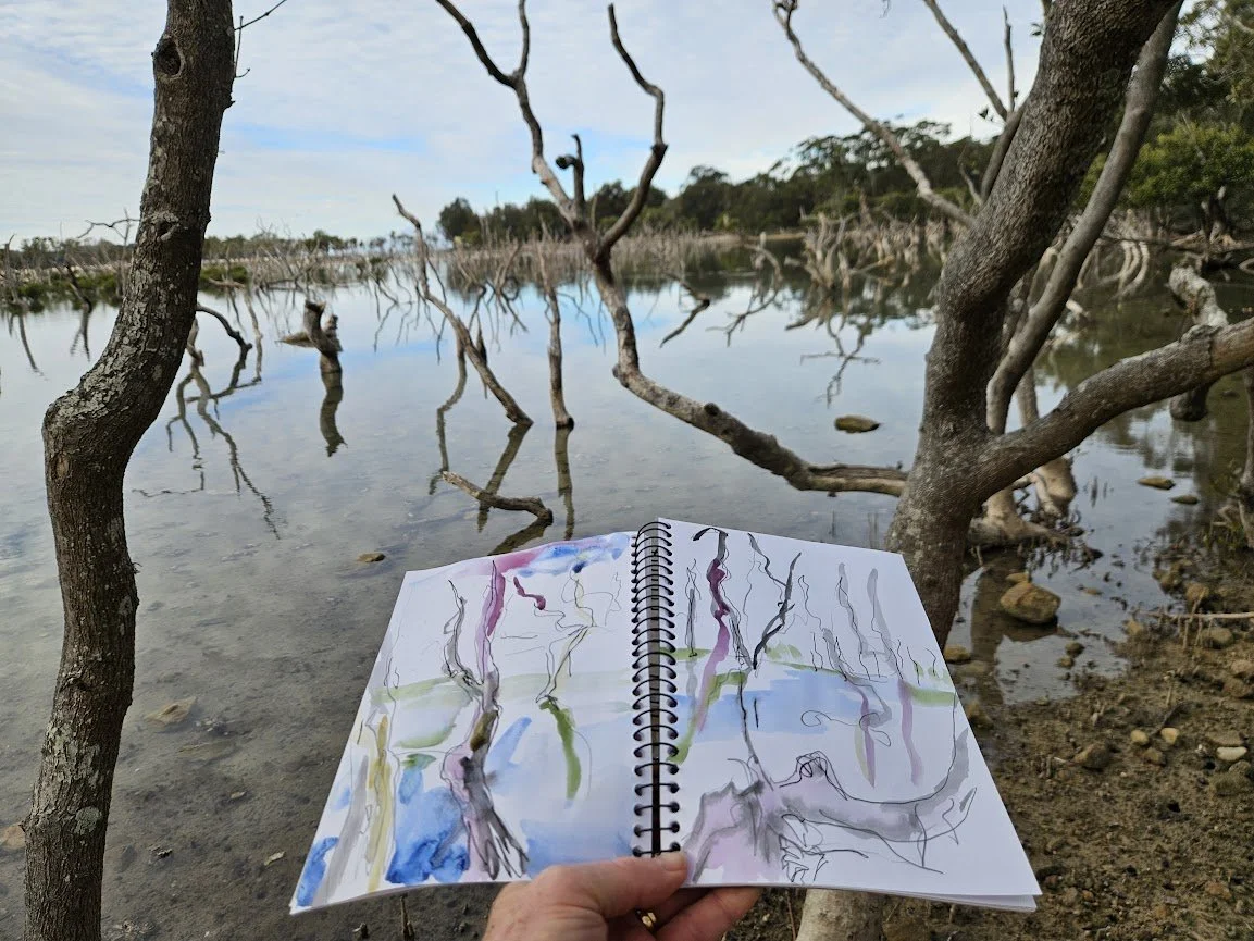 A hand holding an open sketchbook with watercolor drawings of tree branches, in front of a lake with partially submerged, leafless tree branches and trees in the background.