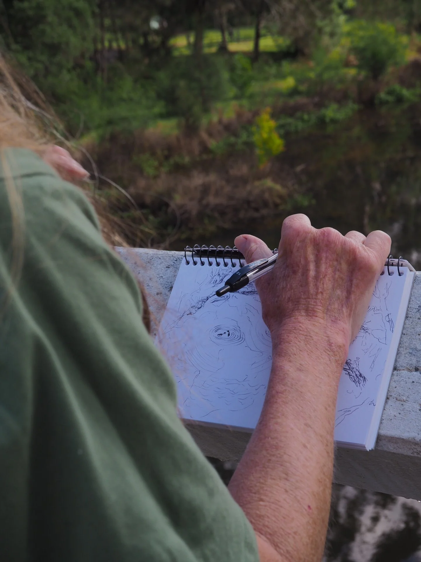 Close-up of an elderly person's hand holding a pen and sketching on a spiral notebook near a river with greenery and trees in the background.