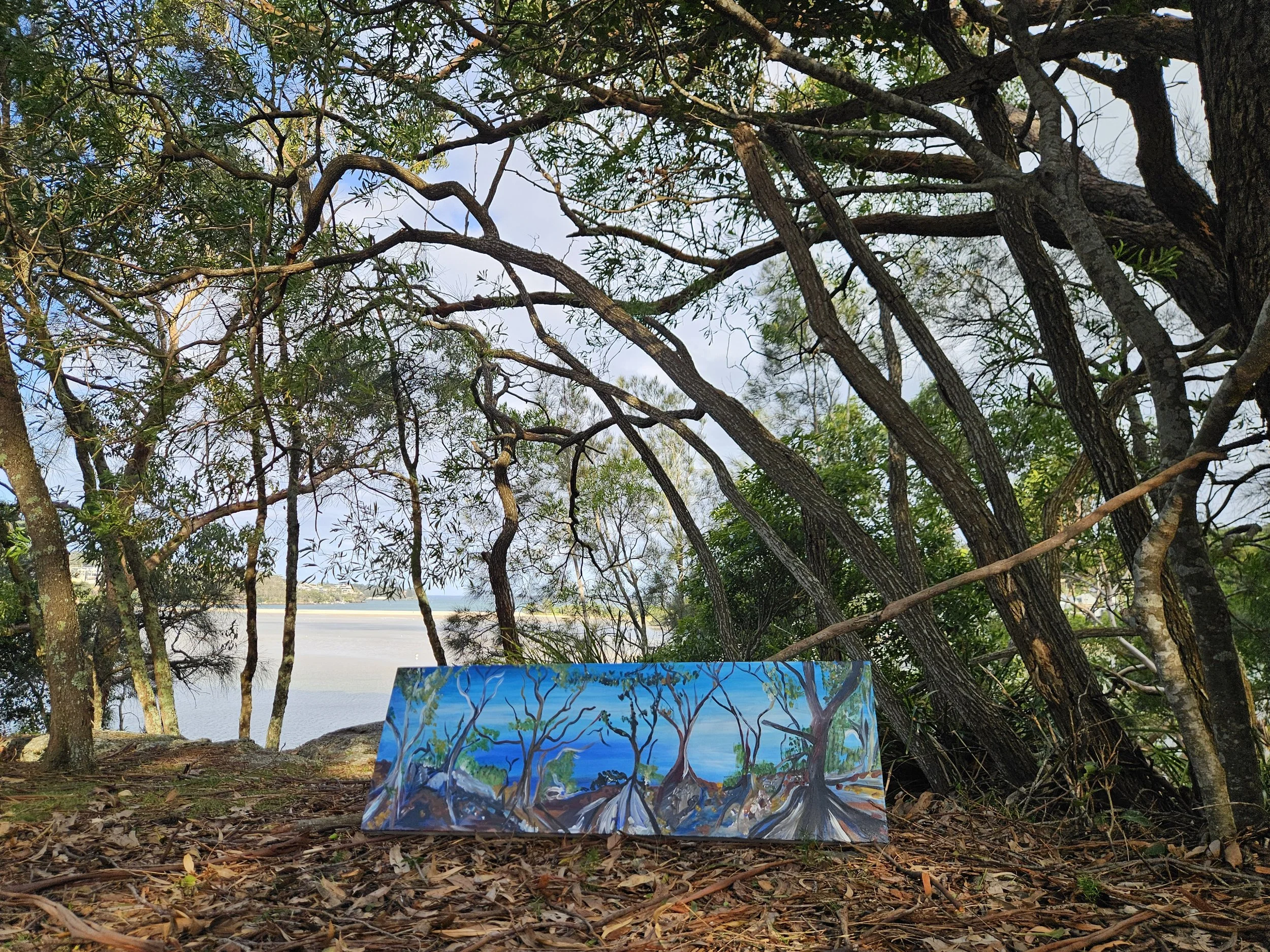 A colorful painting of a landscape with trees, mountains, and a hint of water, placed on the ground among natural trees and branches with green leaves and blue sky in the background.