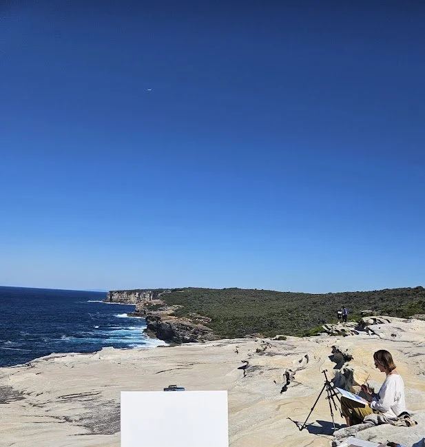 Person painting on rocky coastal landscape with ocean and cliffs under clear blue sky.