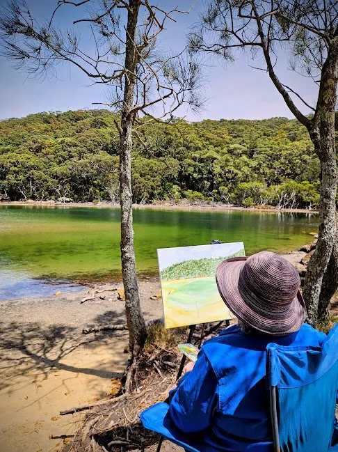 Person sitting on a beach near a lake, painting a landscape on a canvas, wearing a large floppy hat and blue jacket, with trees and hills in the background.