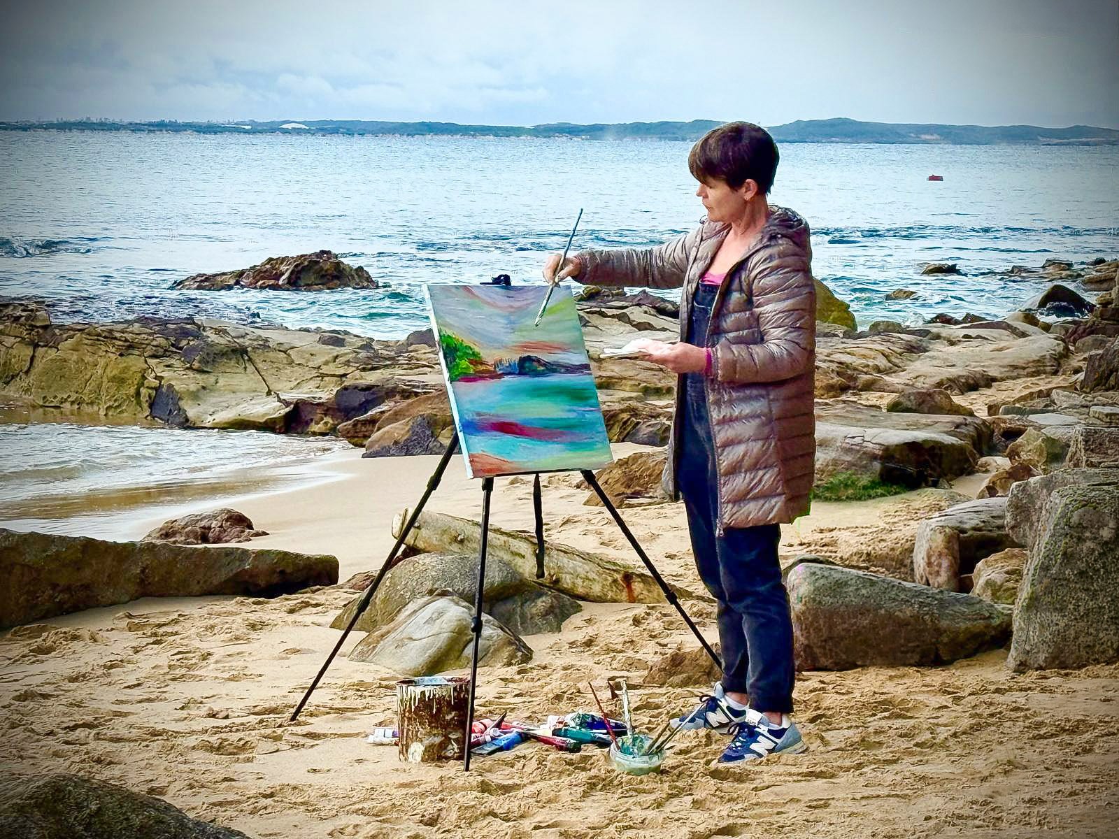 A woman standing on a beach in Bundeena, Royal National Park, painting a landscape painting en plein air.