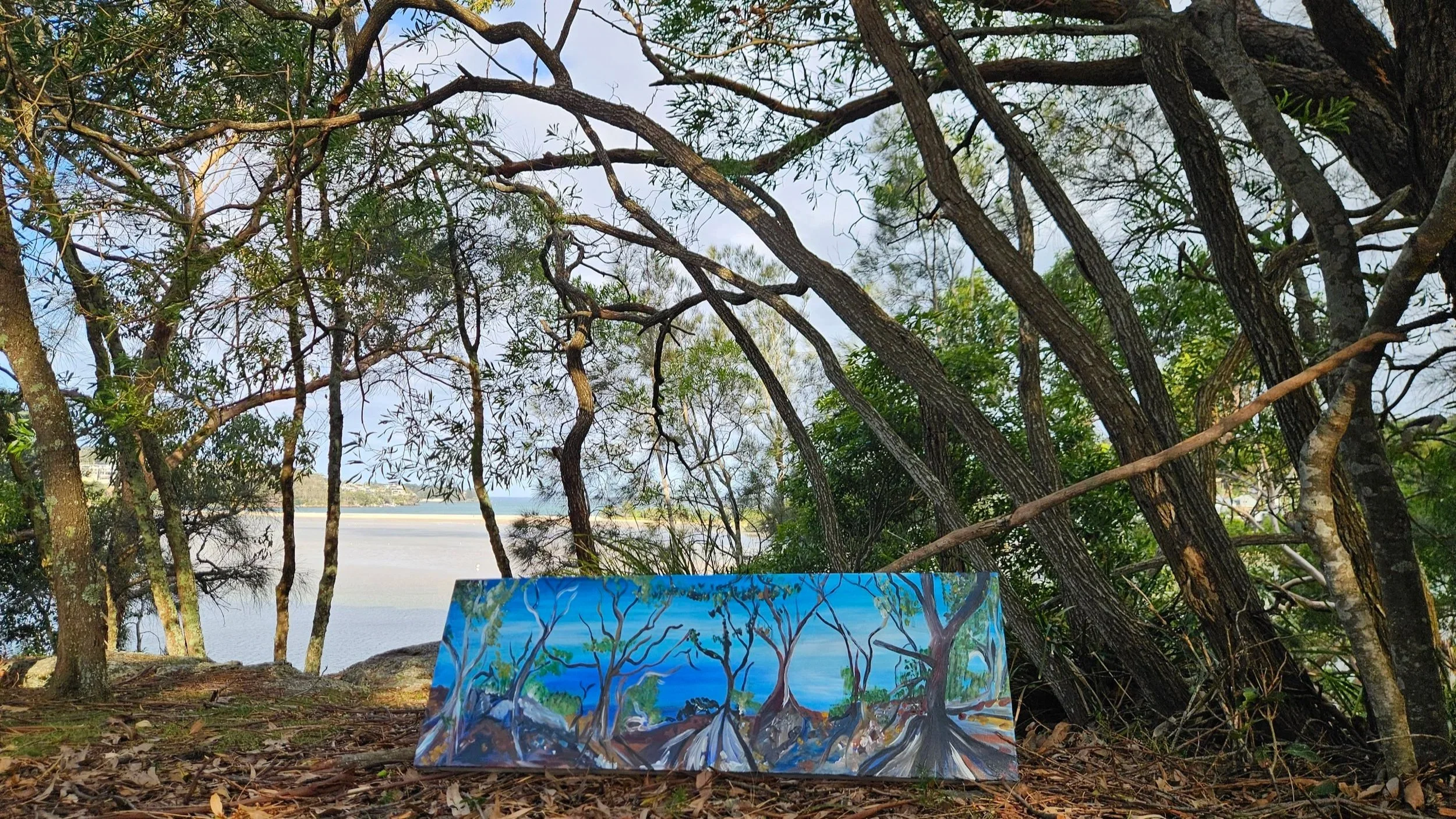 Painting of a tropical landscape with trees, mountains, and water, leaning against a tree trunk outdoors by a body of water.