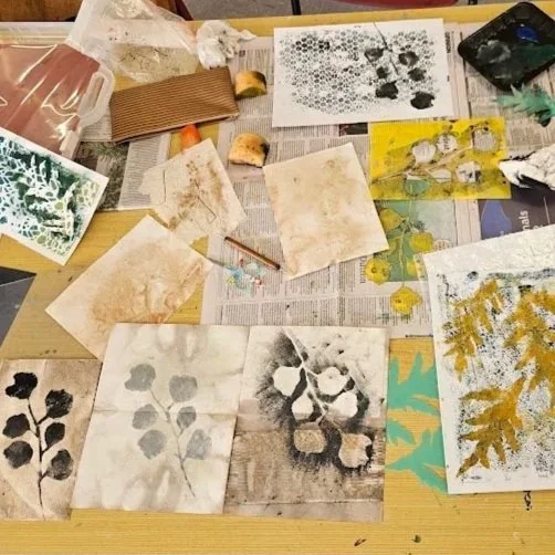 Art supplies and painted papers on a table, including paint palettes, brushes, and paper with leaf and plant patterns.
