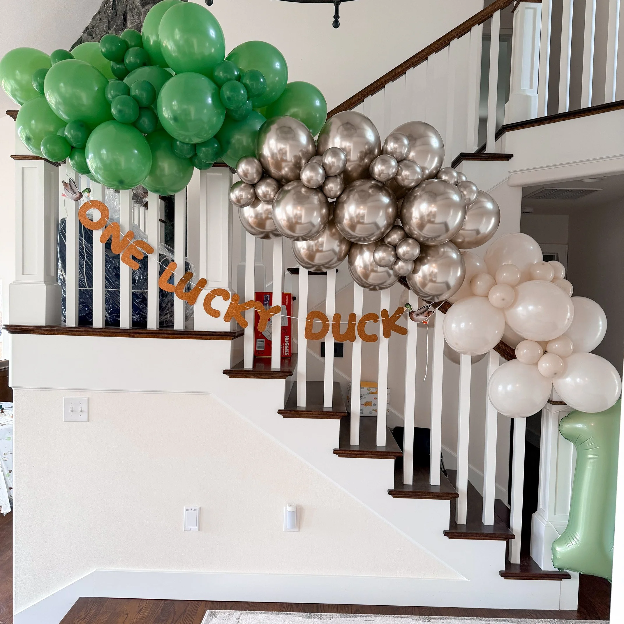 Decorative balloon arrangement on staircase banister with orange banner reading 'One Lucky Duck' and some small animals attached to the banner.