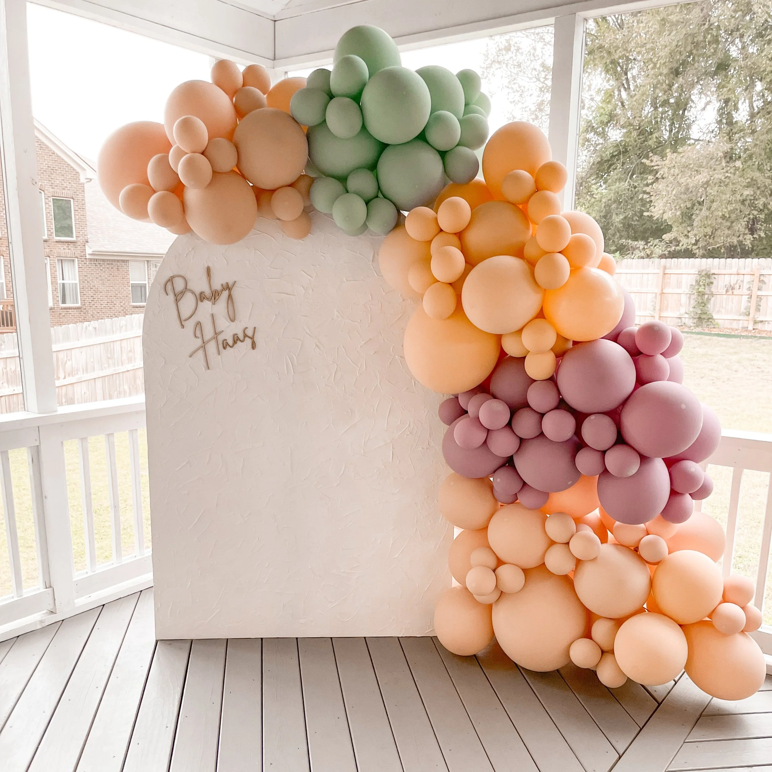 Balloon arch with pastel colors (green, peach, purple, pink) decorates a white textured panel with "Baby Hays" written on it, inside a room with large windows overlooking a backyard.