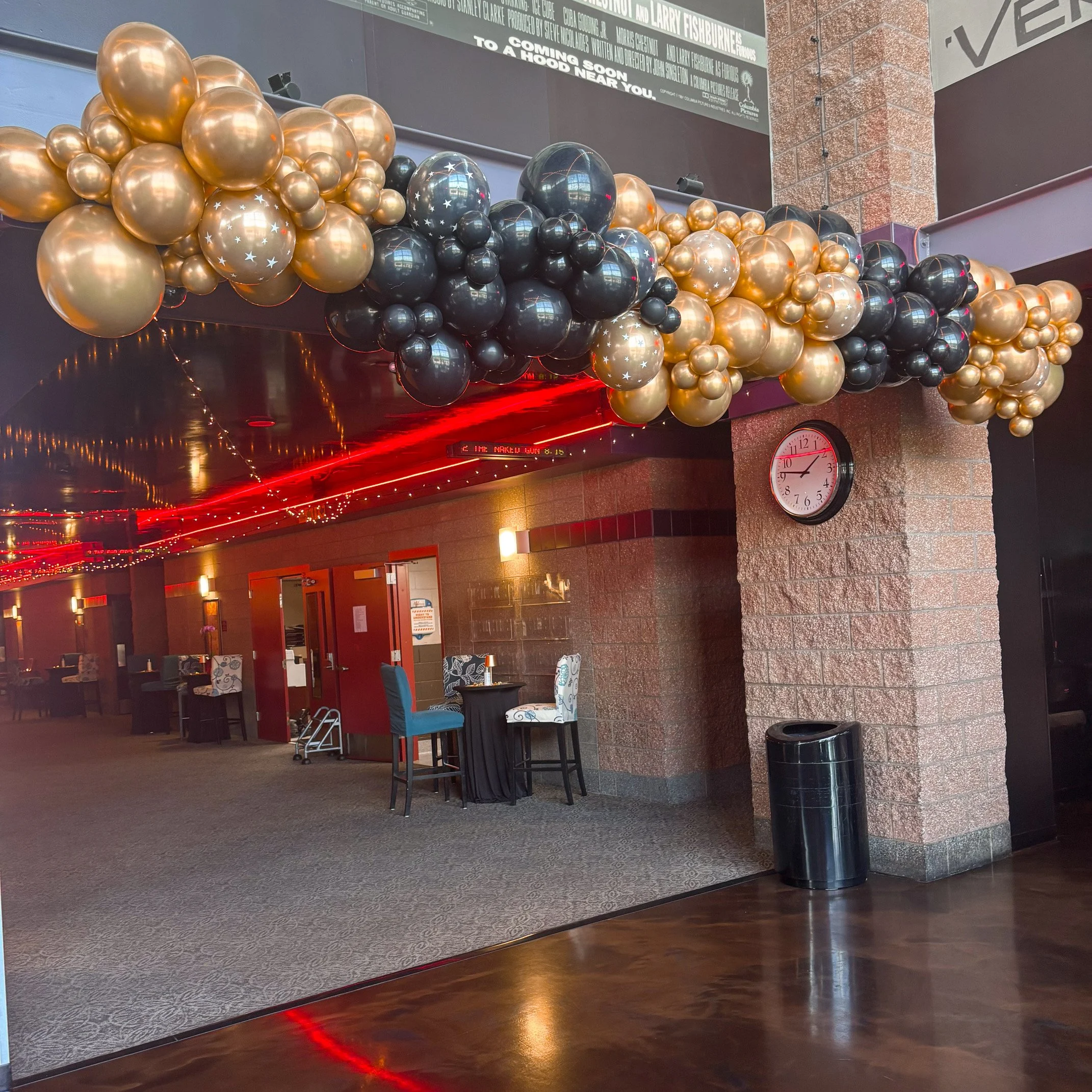 Decorative balloon arrangement with gold, black, and silver balloons hanging from the ceiling in a restaurant or event space.