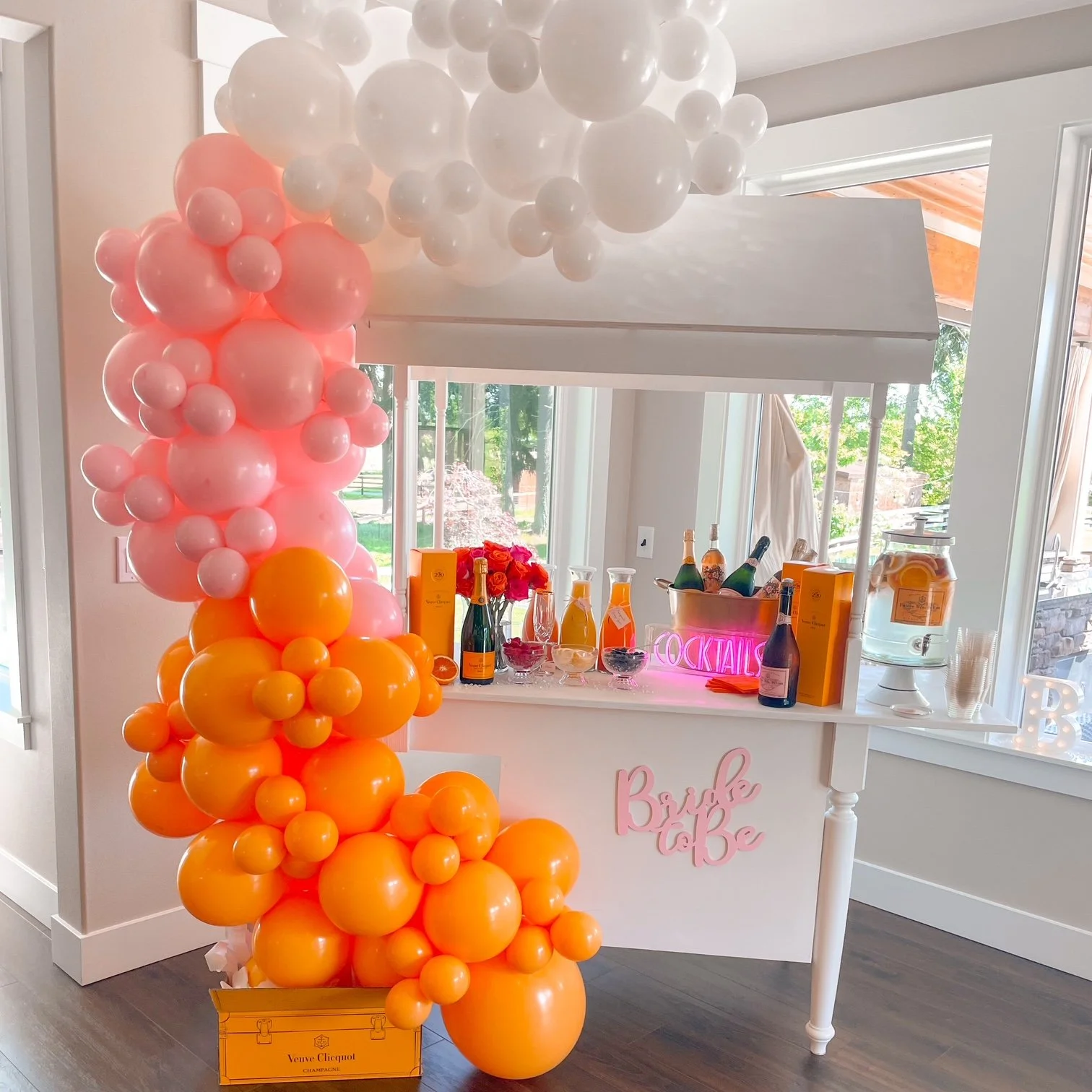 Birthday celebration setup with a balloon cascade of pink, orange, and white balloons, a cocktail bar with drinks, flowers, and lit sign, and a sign that says 'Bride & Be'.