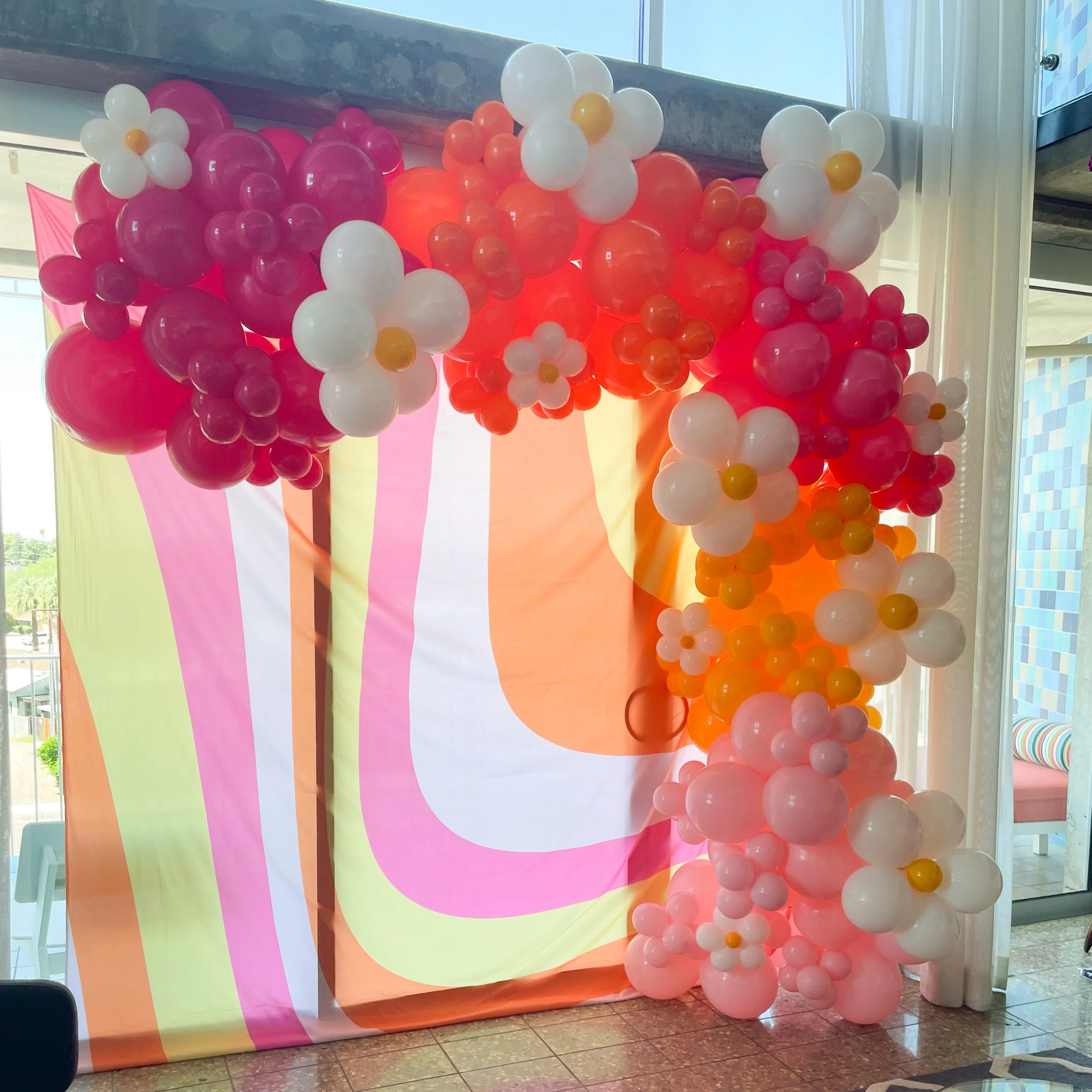 Balloon art display with pink, white, orange, and yellow balloons arranged around a colorful backdrop with pastel stripes.