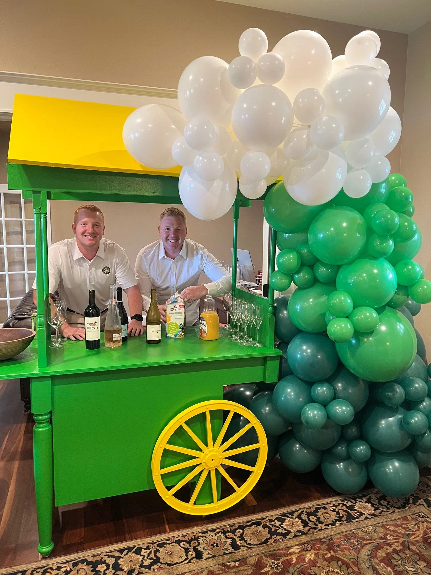 Two men standing behind a green and yellow balloon decorated bar cart with white and green balloons attached, set up indoors.