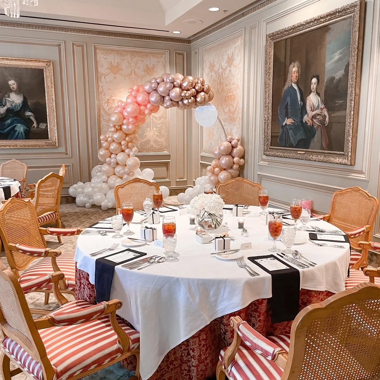 Elegant banquet table set for a celebration, decorated with pink and white balloons forming an arch, with pastel-colored chairs, floral centerpiece, and framed classical portraits on the wall.