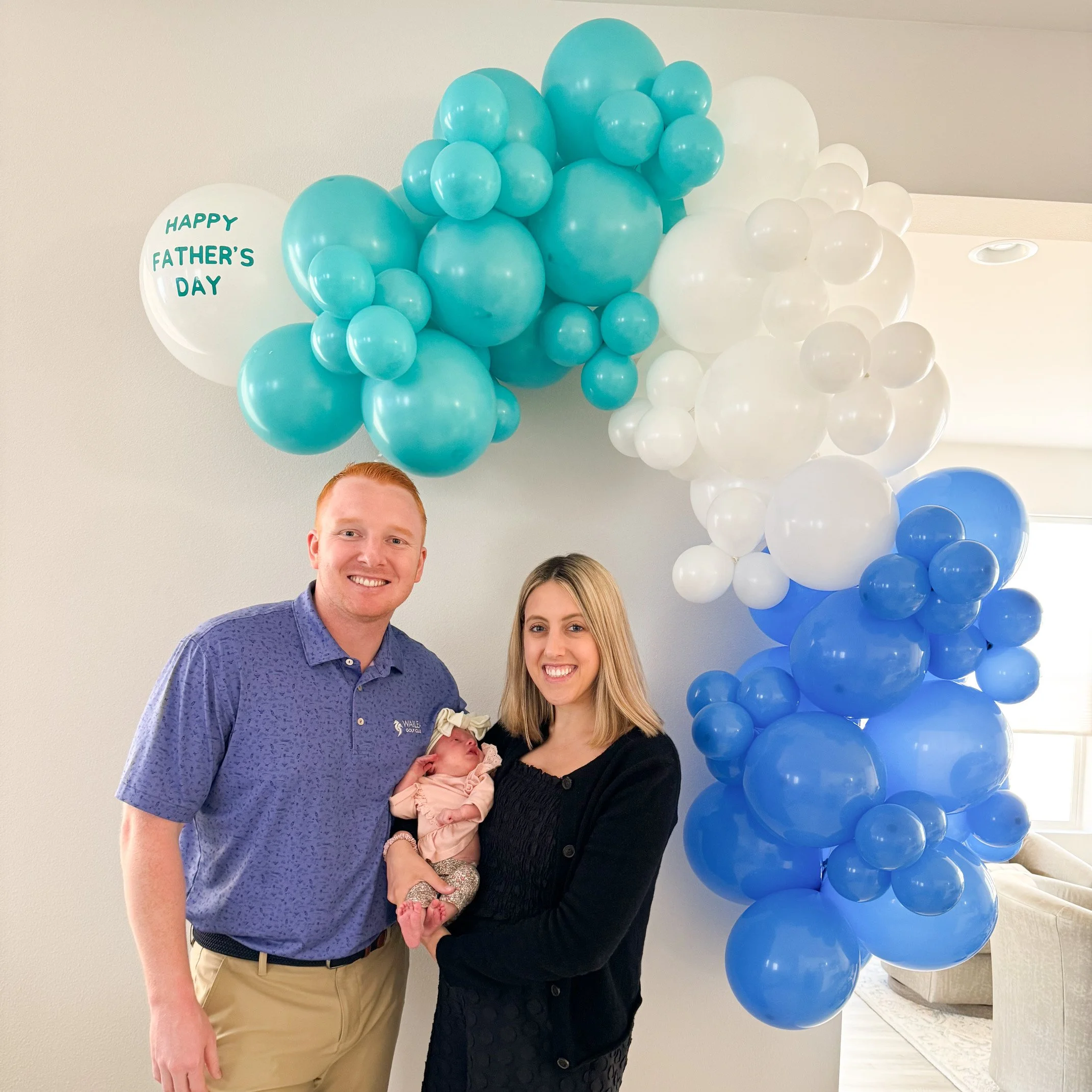 A couple holding a newborn baby standing in front of a Father's Day balloon decoration with blue, white, and turquoise balloons, and a white balloon with green text that says 'HAPPY FATHER'S DAY'.