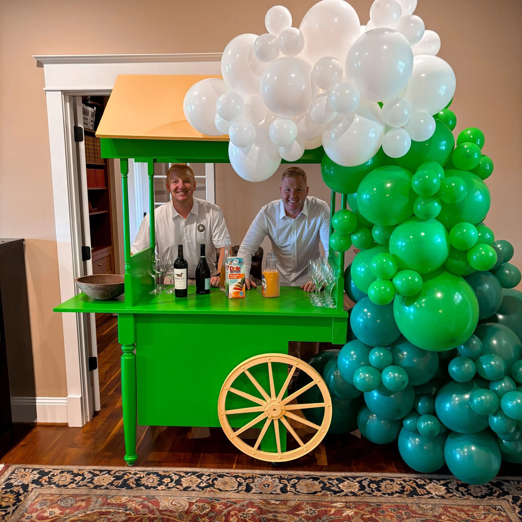 Two men in white shirts behind a small green cart with balloons and drinks, resembling a festive food or drink stand, in a room with hardwood floor and a patterned rug.